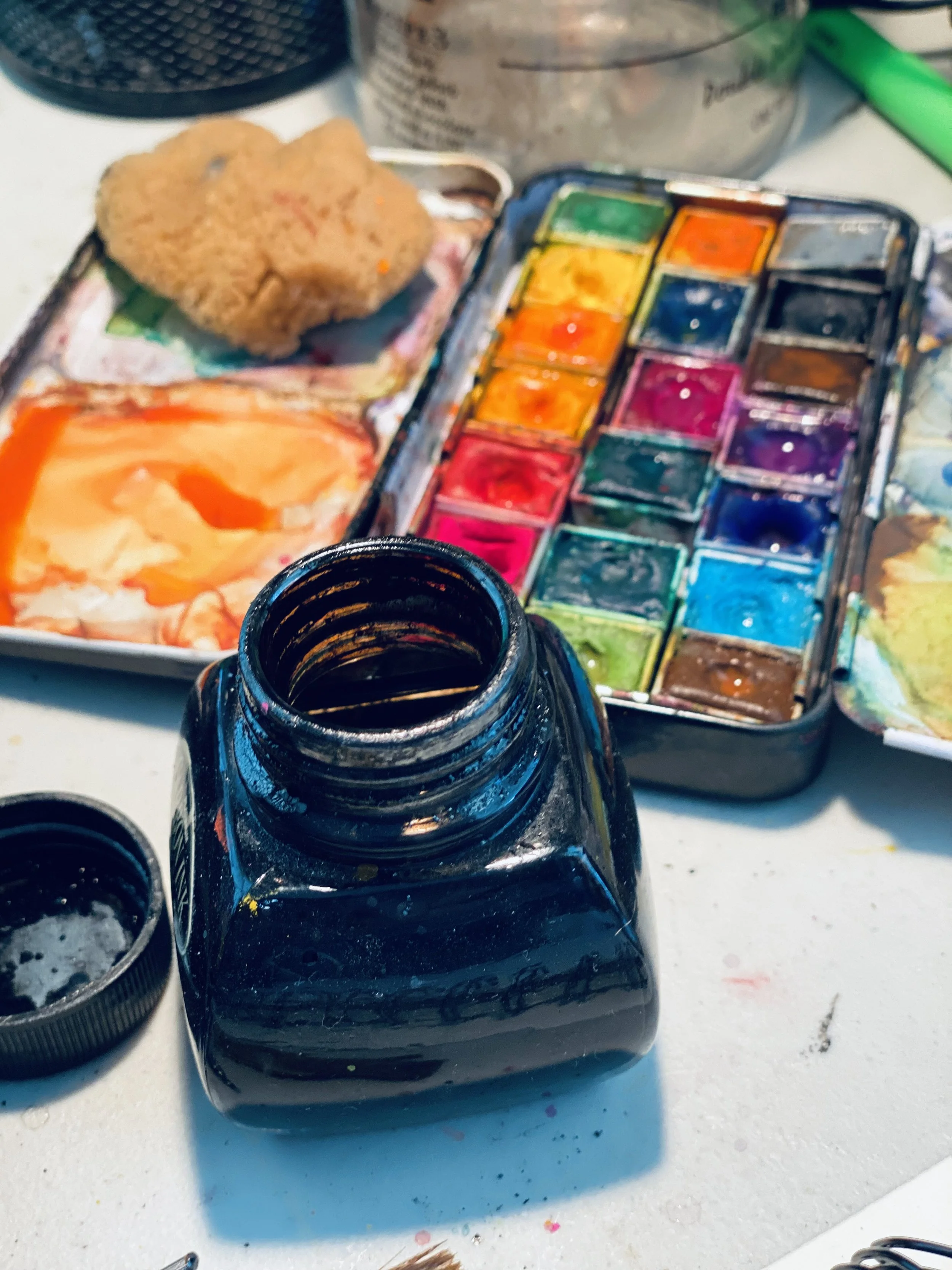 Open black bottle of watercolor paint surrounded by additional paint bottles and mixing tray with various colors, including green, yellow, orange, blue, purple, and red. An out-of-focus canvas or paper with paint swatches is in the background.