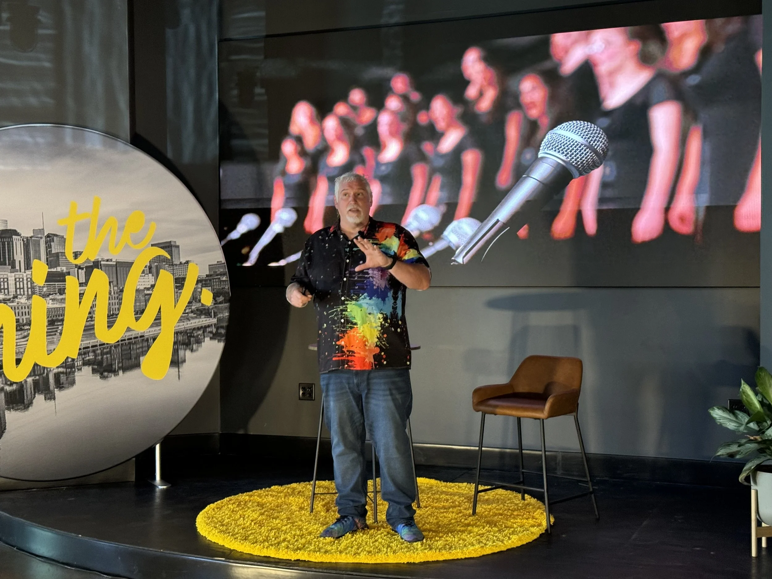 Mike Brennan giving a presentation on a stage, standing on a yellow rug in front of a large screen showing a group of people with microphones. There is a decorative cityscape with yellow writing on the left and some plants on the right.