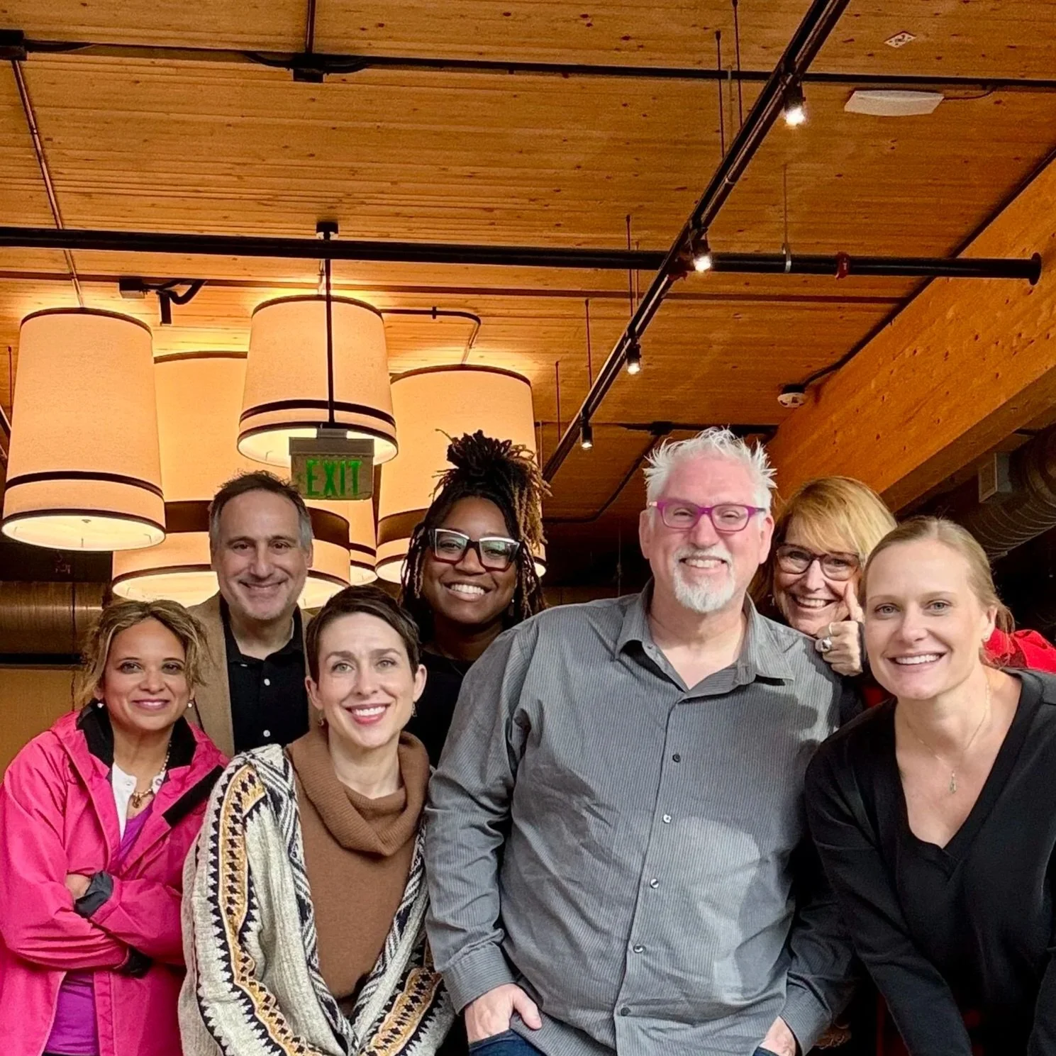 Mike Brennan with a group of seven smiling people posing inside a wooden-paneled room with hanging lamps and an exit sign overhead.