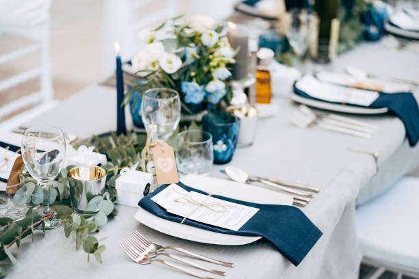  A square plate with a blue cloth towel, knives and forks next to the plate. Flower composition with eucalyptus leaves in the center of the table and burning candles. 