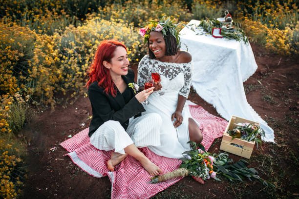  Lesbian wedding picnic party. Two brides sitting down on the blanket, making a toast with champagne 