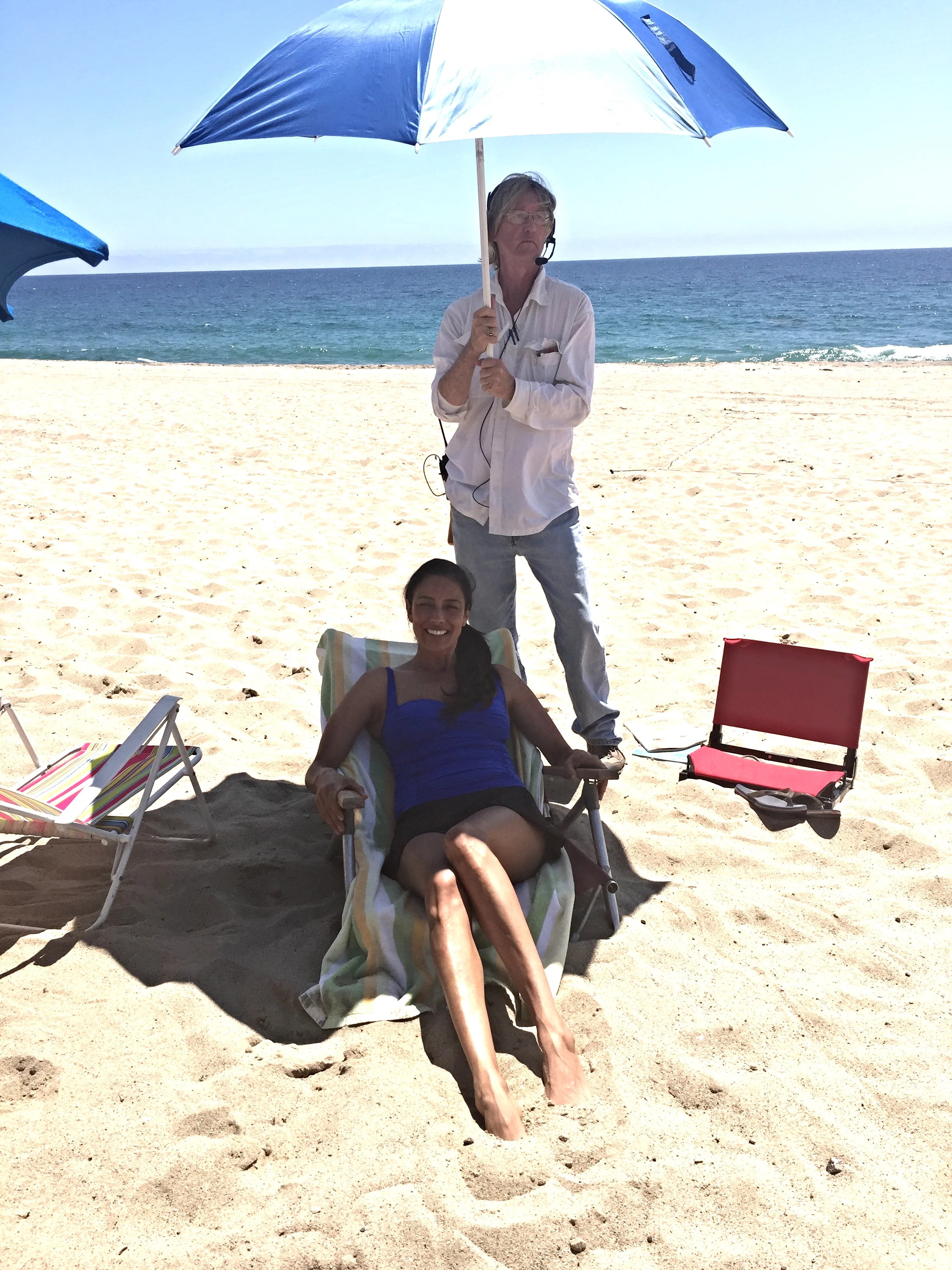 A woman relaxing on a beach chair under an umbrella with a man standing behind her holding another umbrella, both near the shoreline with the ocean in the background.