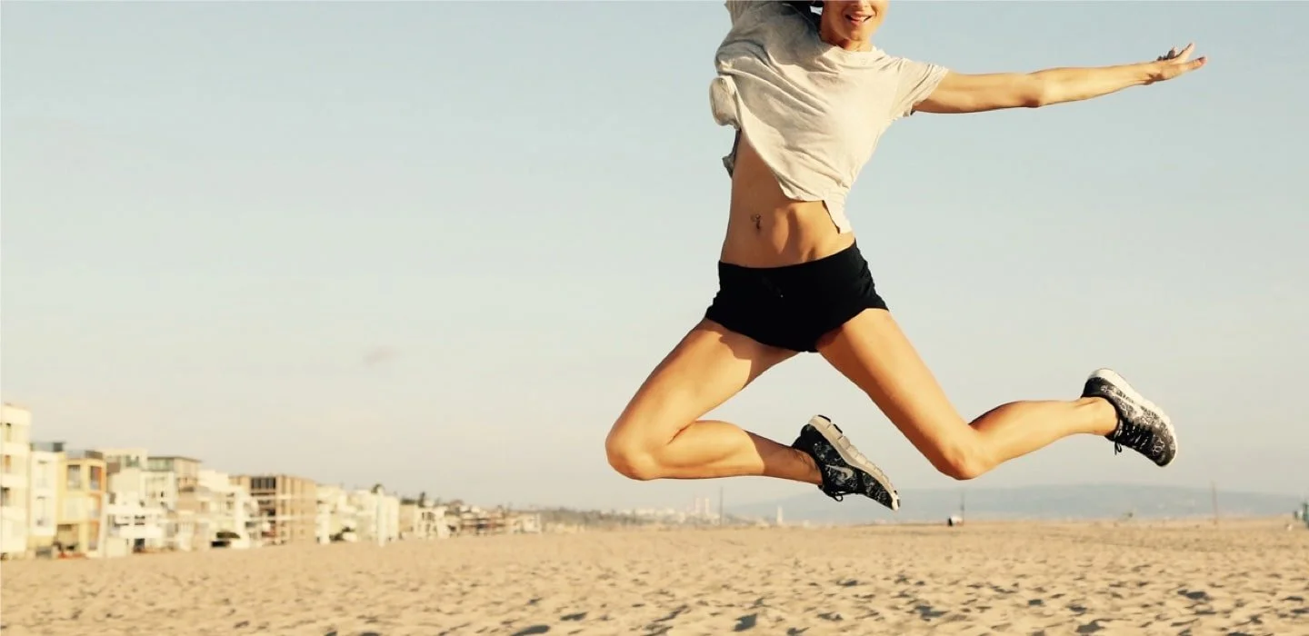 A woman is mid-air, jumping on a beach, with stretch marks visible on her abdomen, wearing a beige t-shirt, black shorts, and athletic shoes.