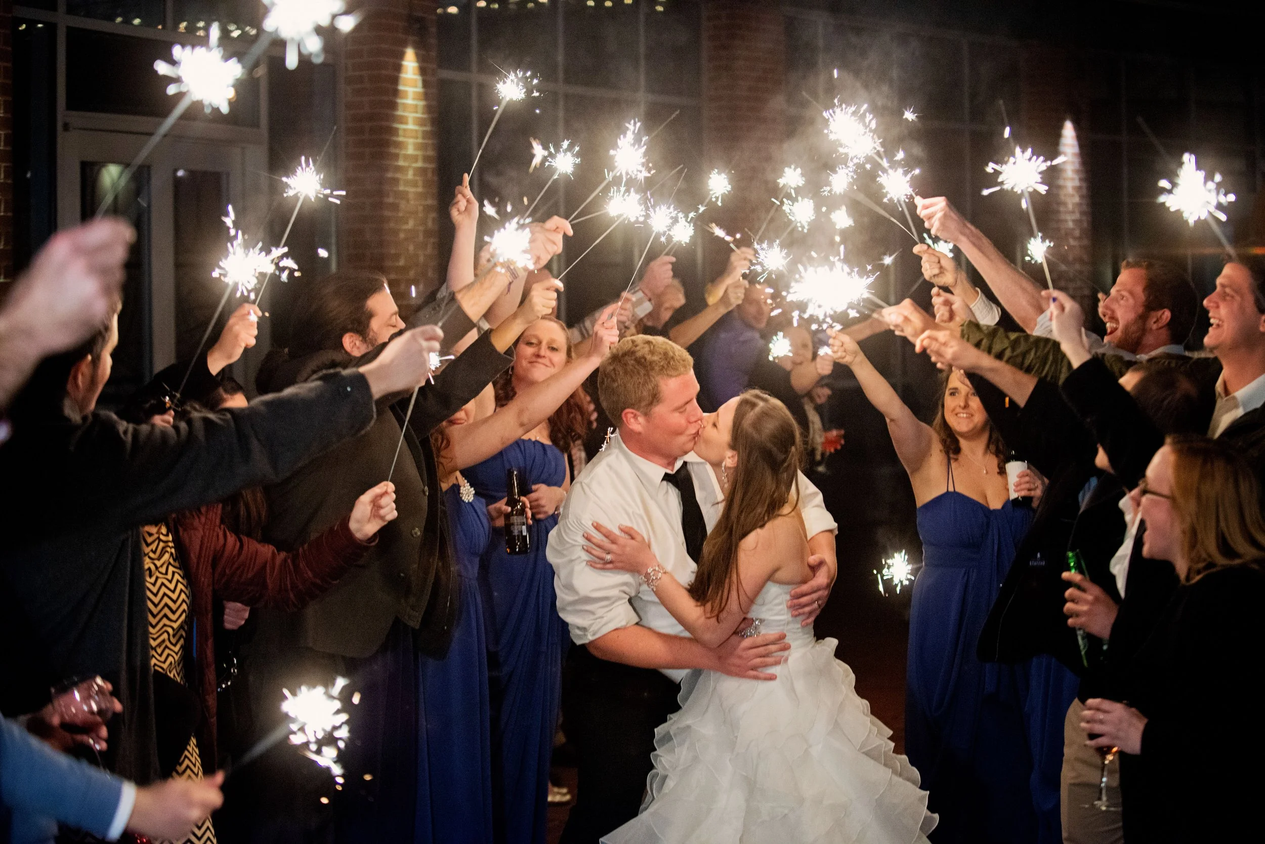 Newlyweds kissing during a joyful sparkler exit surrounded by friends and family