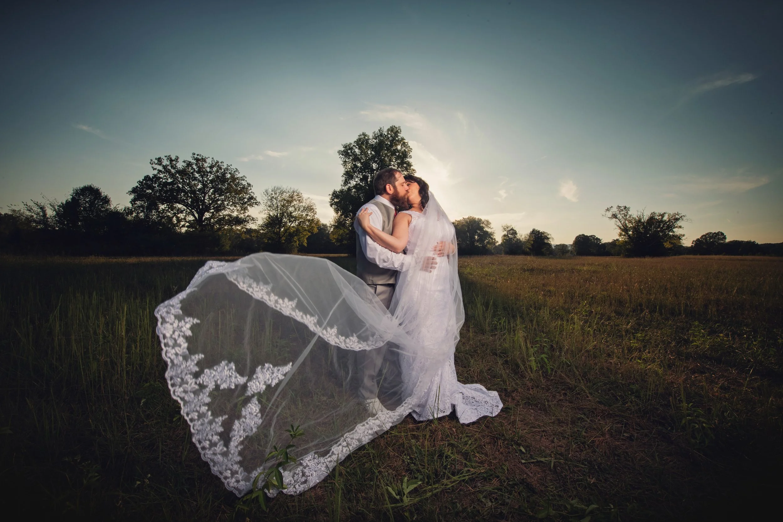 Newlyweds embracing in an open field with the bride's veil caught in the breeze