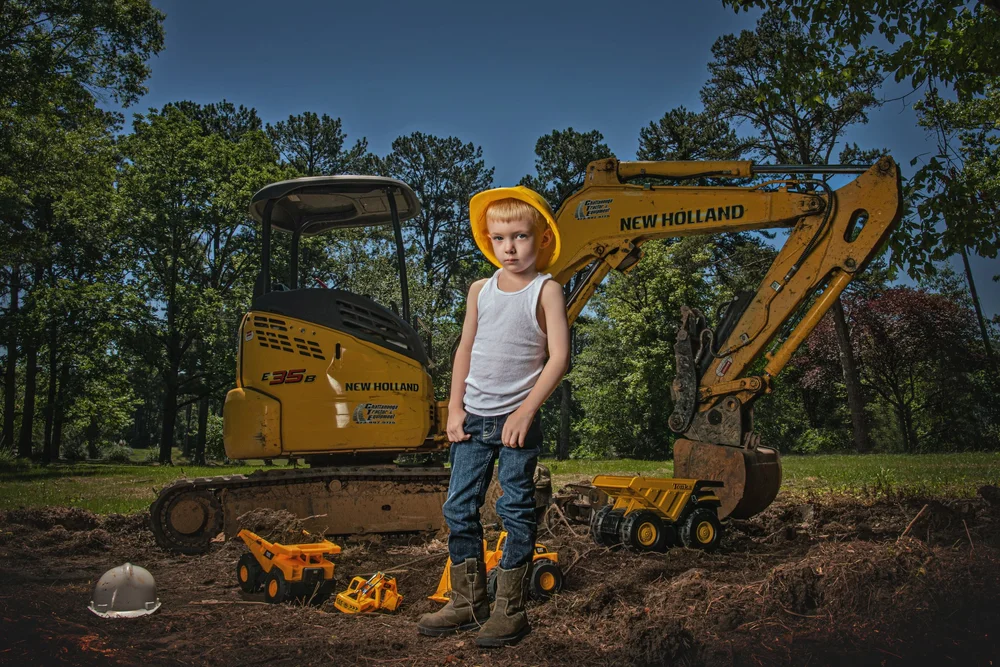 Child standing beside construction equipment and toy trucks
