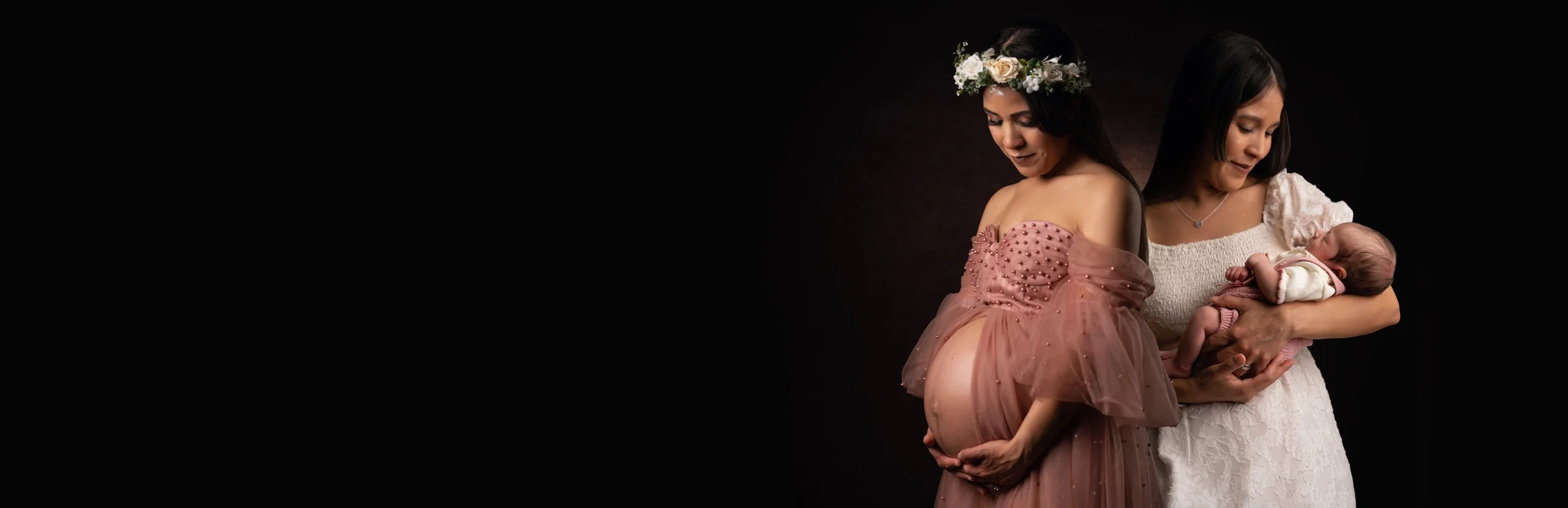 A pregnant woman in a pink dress with a flower crown cradles her belly, accompanied by another woman in a white dress holding a baby, against a black background.