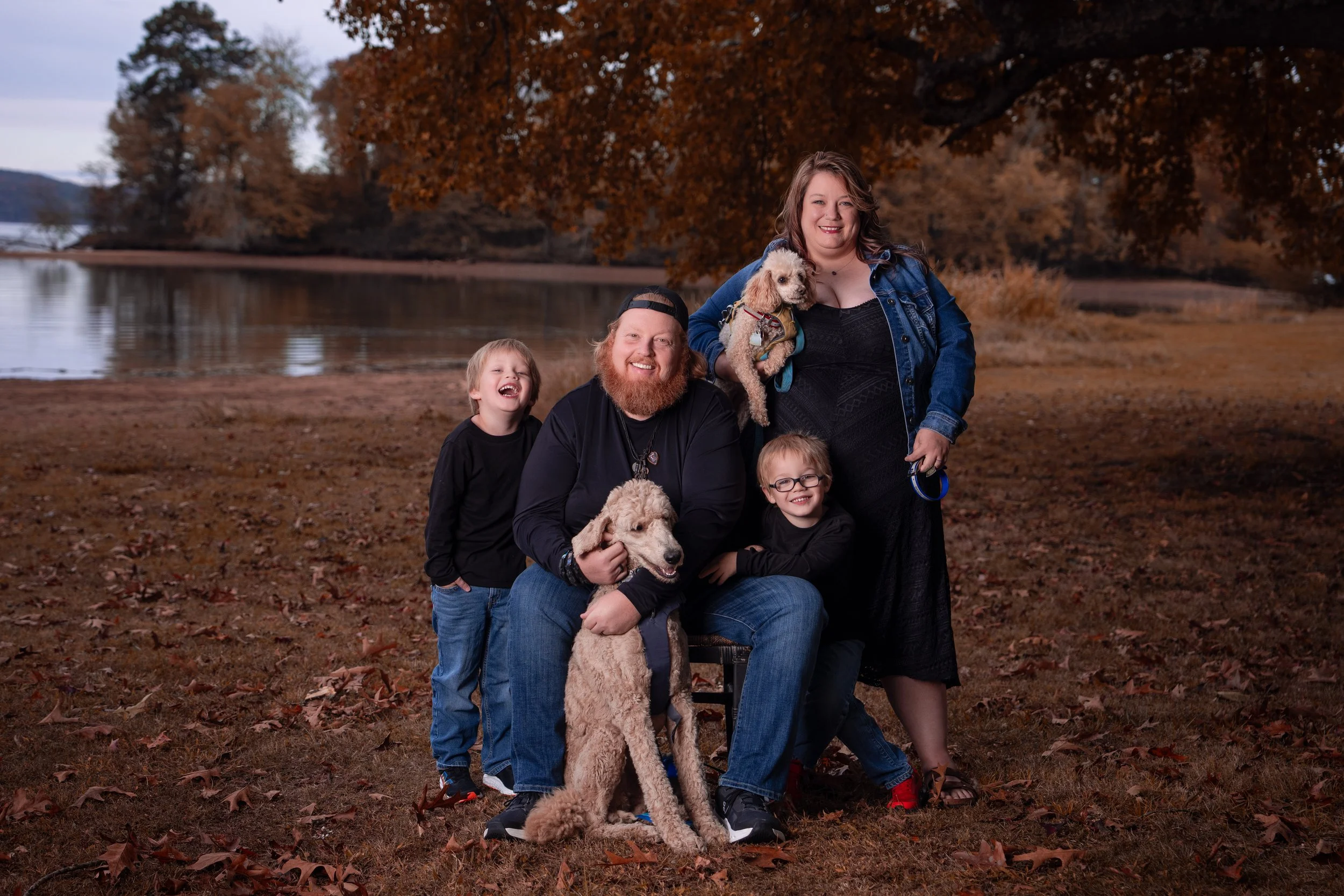 A family of five and two dogs outdoors in fall, with trees and a lake in the background, smiling.