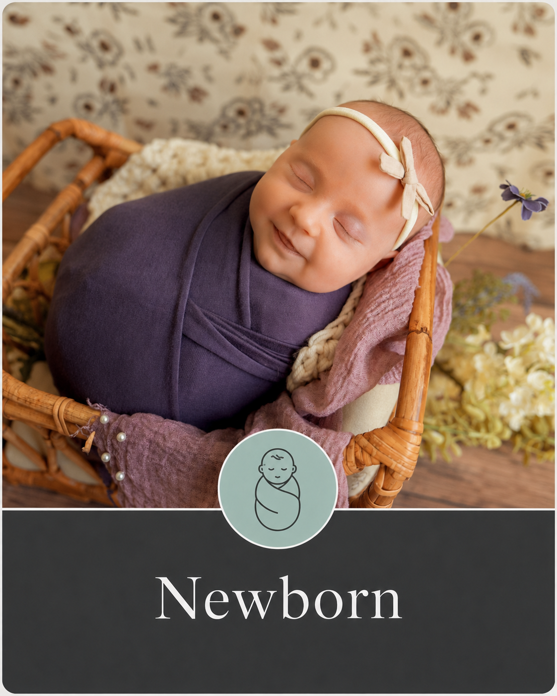 A smiling newborn baby wrapped in a purple blanket, wearing a cream-colored headband with a bow, lying in a rattan basket filled with soft pink and cream fabrics, surrounded by flowers.