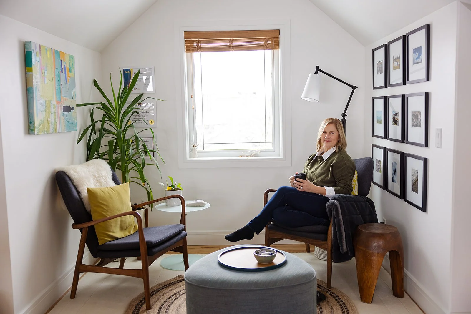 A woman sitting in a cozy living room corner with a cup in her hands, surrounded by modern artwork, a large potted plant, and a gallery wall of framed photos, illuminated by natural light from a window.