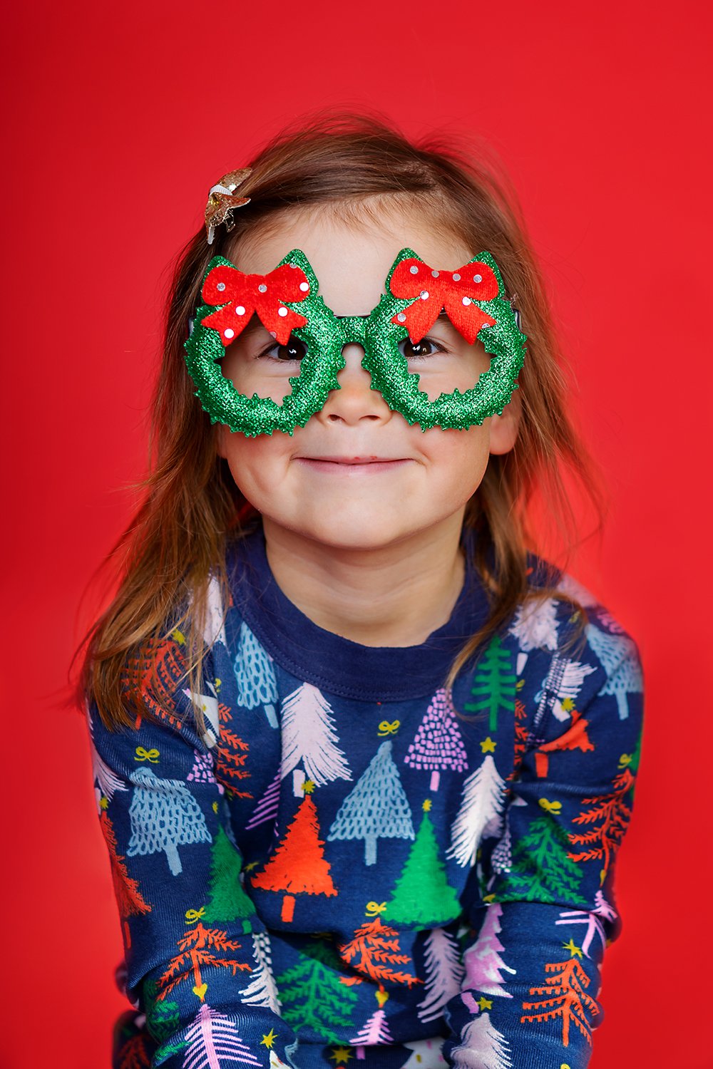 A young girl wearing large, glittery green Christmas wreath glasses with red bows, smiling against a red background, dressed in a colorful Christmas tree patterned sweater.