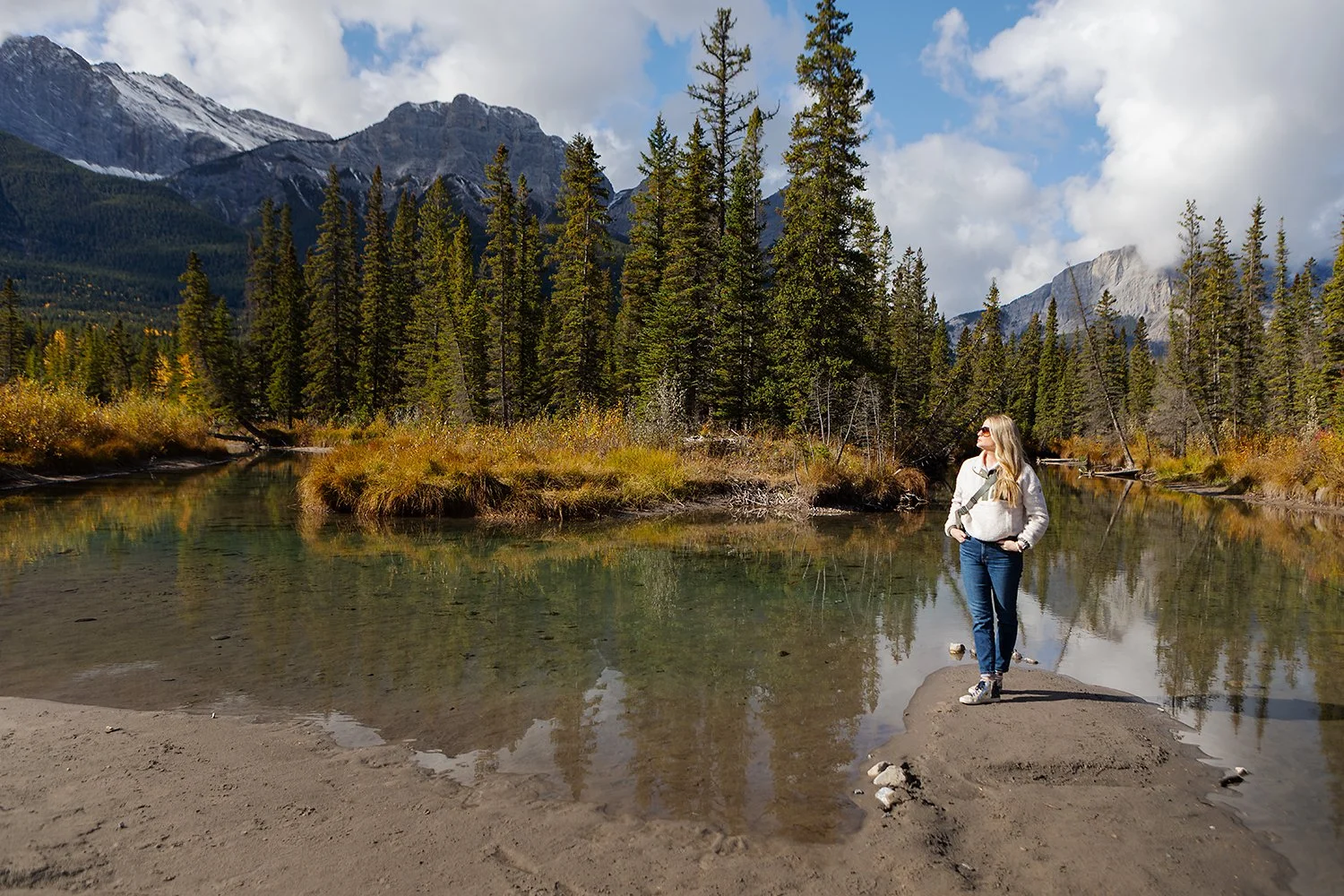 A woman standing on a small sandy islet in a calm river with a forested mountain landscape in the background.