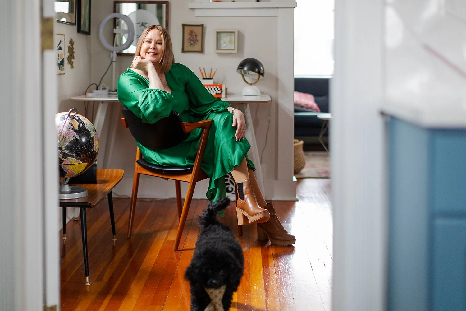 Woman with shoulder-length blonde hair sitting on a wooden chair in a cozy, decorated room, smiling, wearing a shiny green dress and brown boots. A black and gray dog with curly fur walks toward the camera on a wooden floor. The room has framed artwo