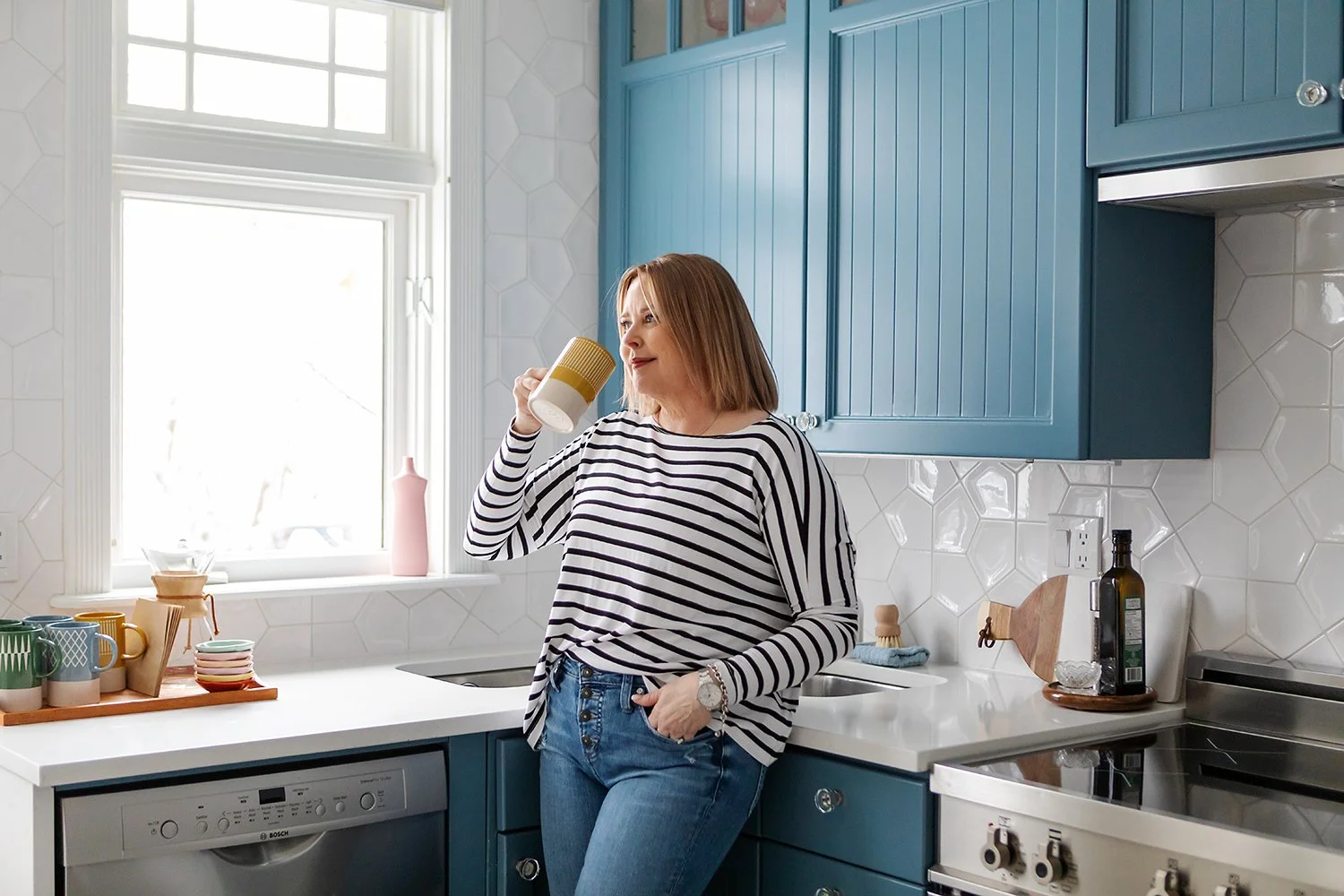 A woman standing in a bright kitchen, drinking from a yellow and white mug, wearing a striped long-sleeve shirt and blue jeans.