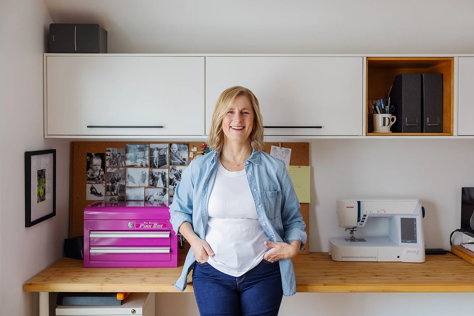 A woman with blonde hair smiling while standing in a craft room with a sewing machine, photos and supplies behind her.