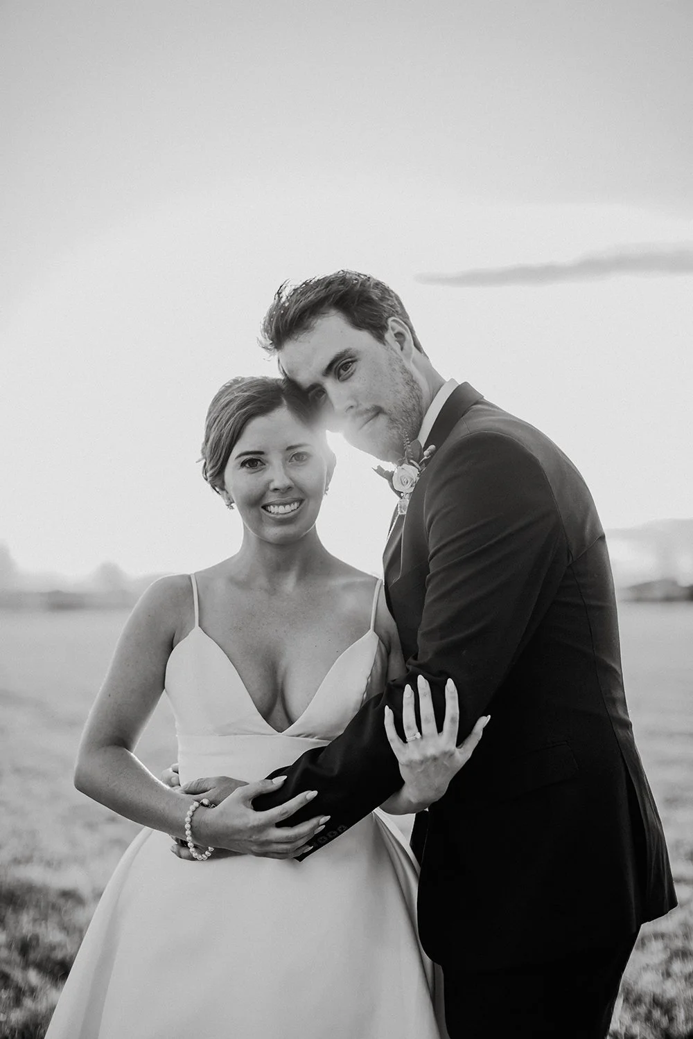 Black and white photo of a bride and groom embracing outdoors at sunset, both smiling at the camera.