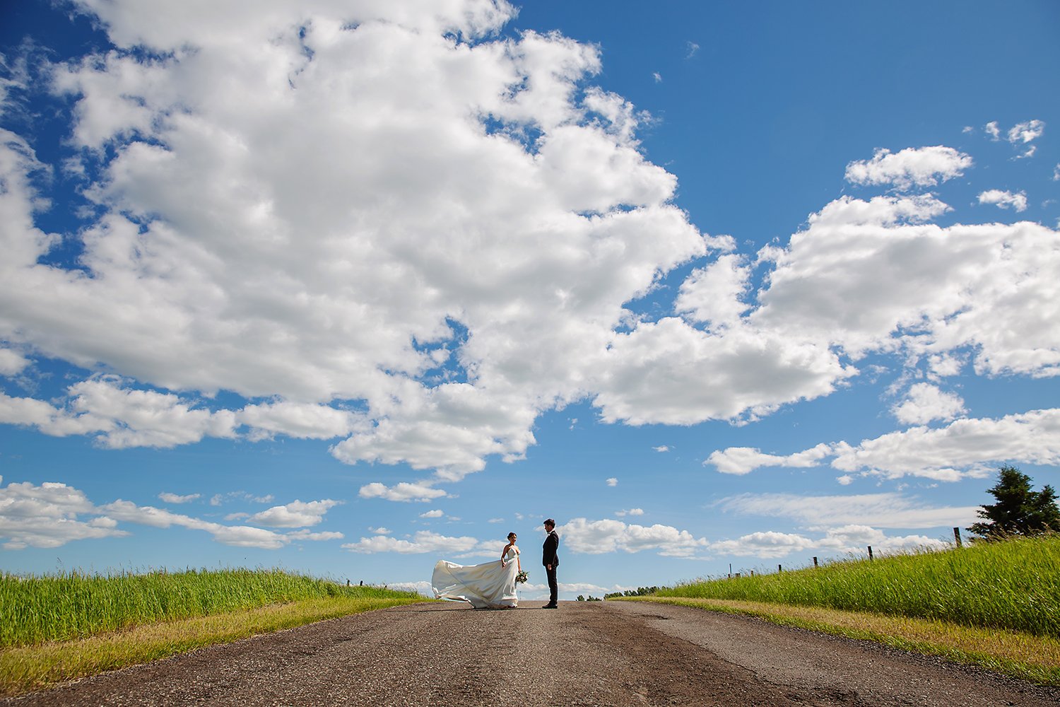 A bride and groom standing on a rural road under a bright blue sky with scattered white clouds, surrounded by green fields.