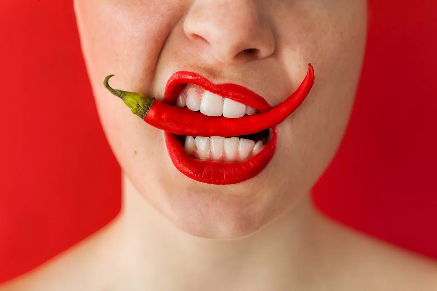 A close-up of a woman's face with bright red lipstick, holding a red chili pepper in her mouth like a smile, against a red background.