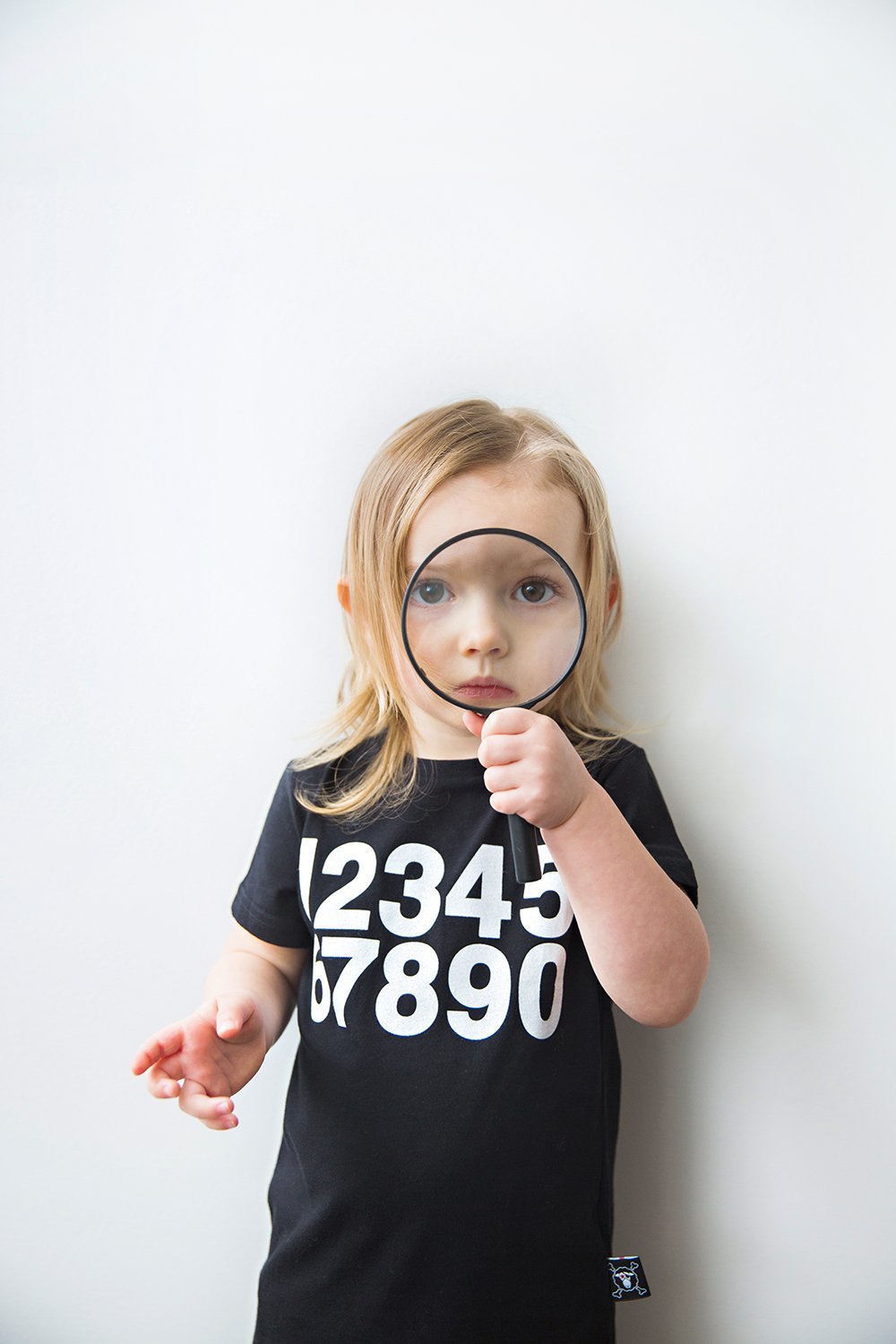Young girl with blonde hair looking through a magnifying glass, wearing a black T-shirt with white numbers from 1 to 9.