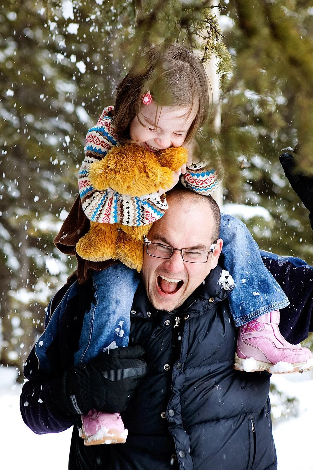 A man carrying a young girl on his shoulders, playing in the snow outdoors, both smiling and enjoying the winter weather.
