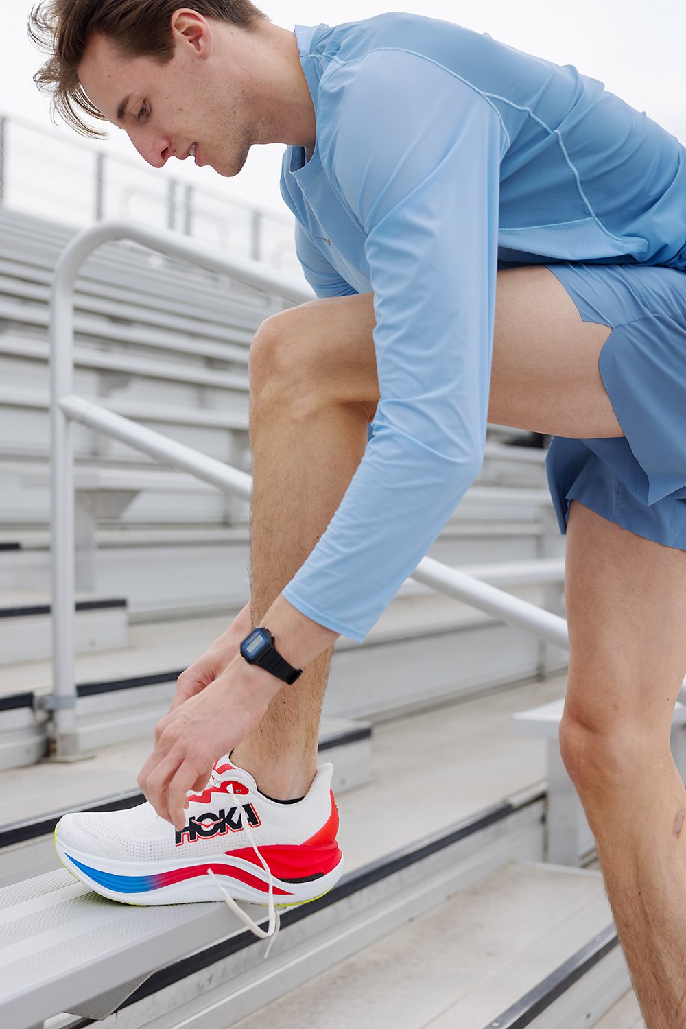 Young male athlete in blue athletic wear tying his white running shoe with red and black details on bleacher steps.