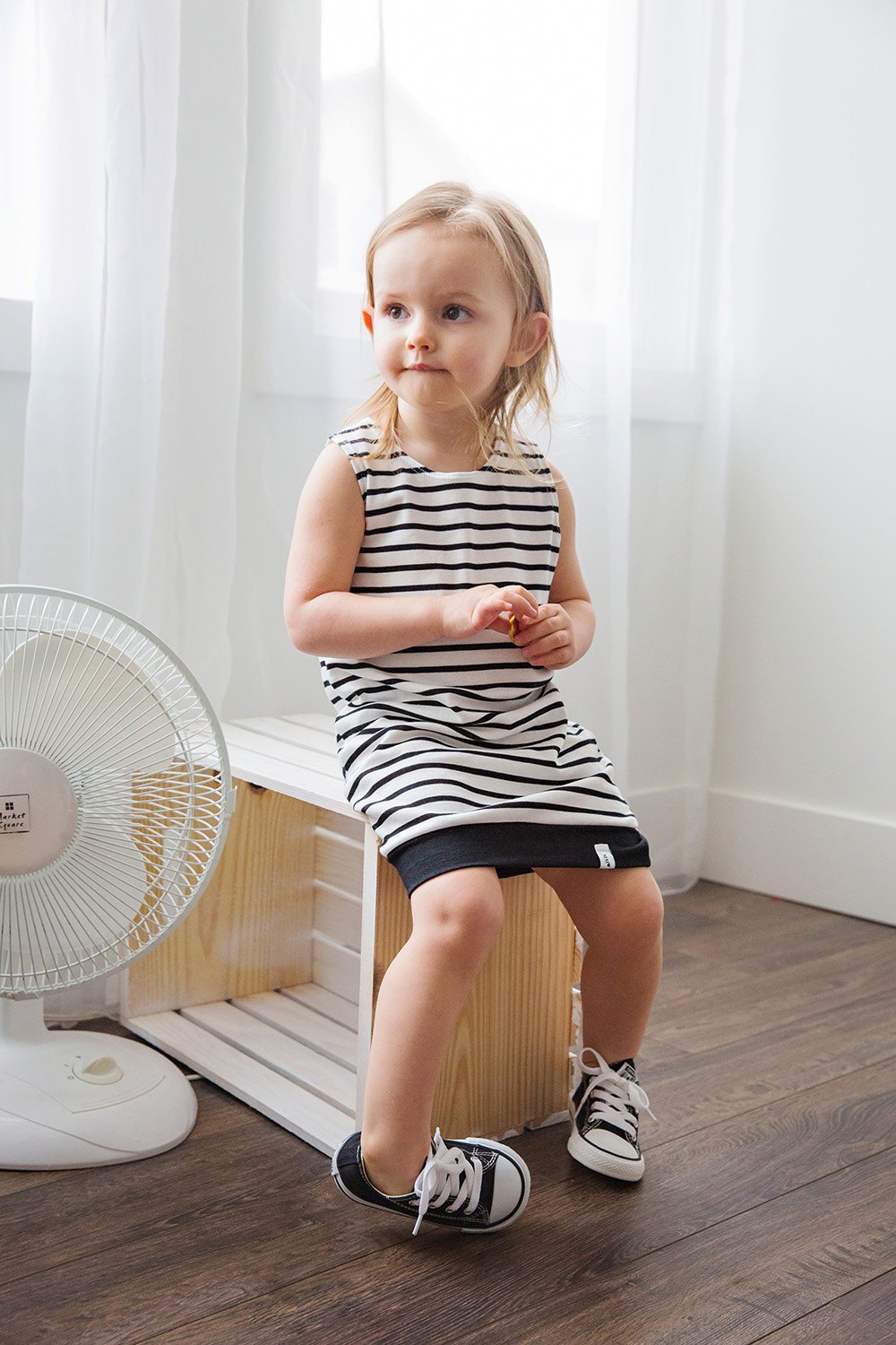 A young girl with blonde hair and blue eyes sitting on a wooden bench in a bright room with white curtains. She is wearing a black and white striped dress and black sneakers. There is a white fan beside her and hardwood flooring.