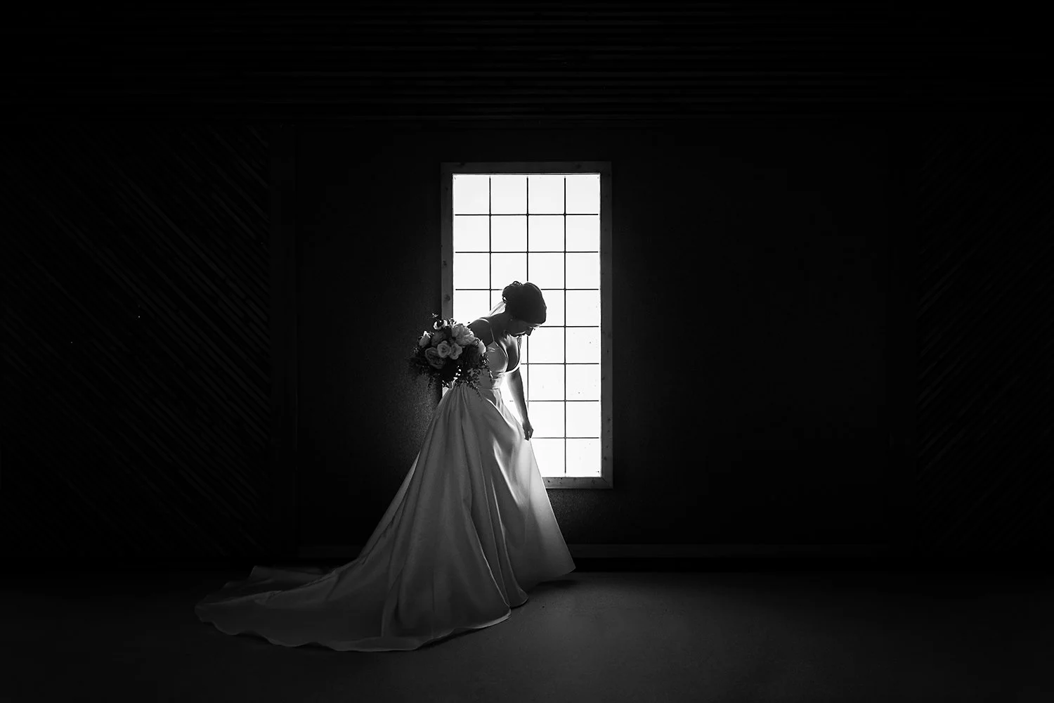 Black and white photo of a bride standing in front of a large window, holding a bouquet of flowers, with her head bowed.