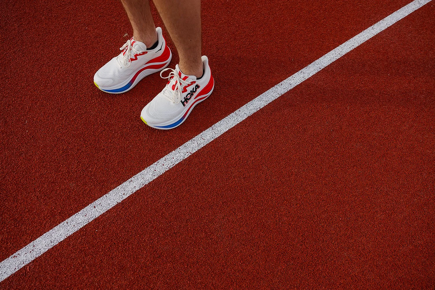 A person wearing white athletic shoes with red, black, and blue accents standing on a red running track next to a white line.