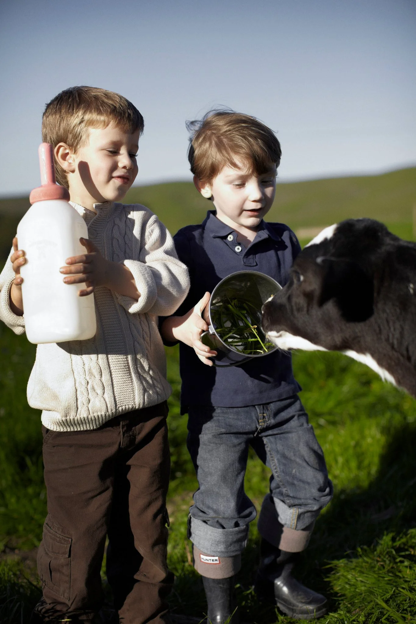   Two boys stood by the wooden fence, their eyes wide with curiosity as they watched a small calf eagerly suckle from a bottle. The scene was peaceful, reminiscent of simpler times spent in the countryside. Nearby, an open copy of the  Tyler Florence