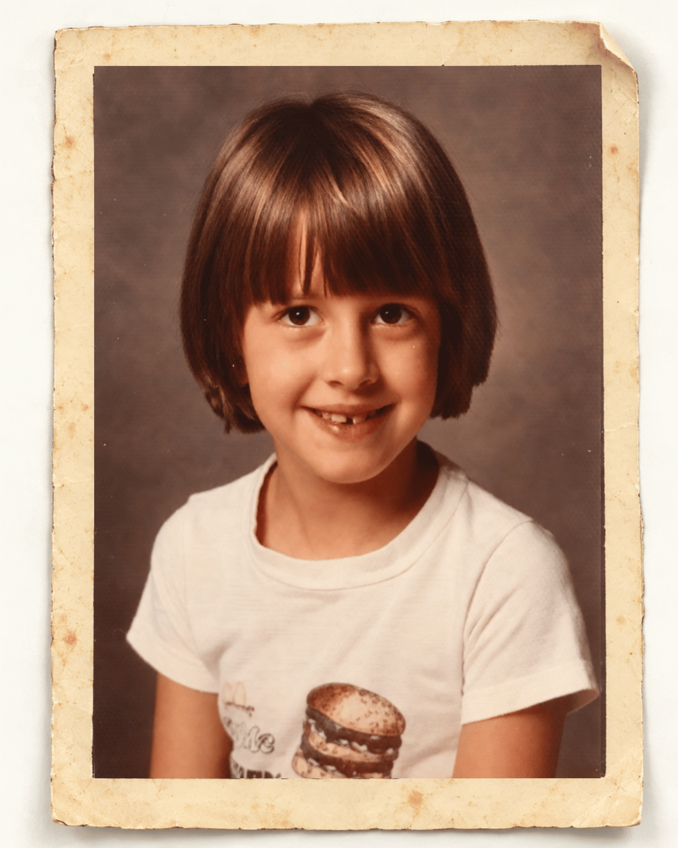 Lauren in second grade, smiling with a missing front tooth and wearing a white T-shirt with a Quarter Pounder graphic, in an aged school-photo paper border.