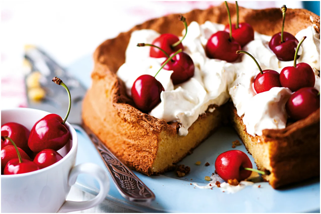 Cherry-topped sponge cake with whipped cream on a blue plate with fresh cherries in a white cup nearby.