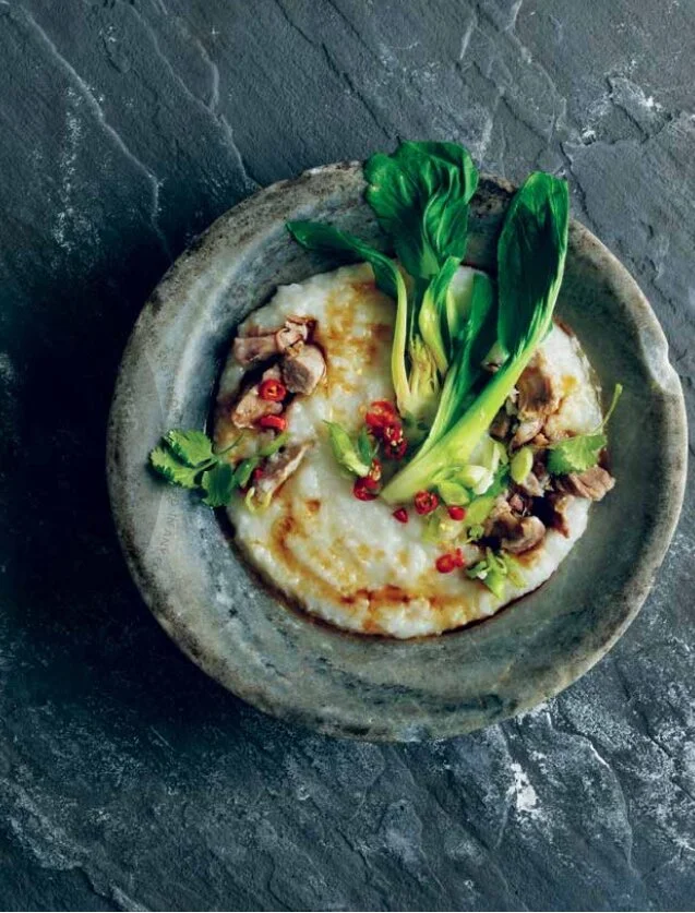A bowl of rice porridge topped with bok choy, chopped meat, red chili slices, and cilantro, served on a dark textured surface.