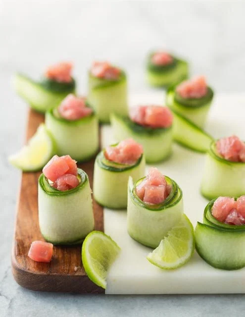 Cucumber rolls filled with chopped raw fish, garnished with lime wedges on a wooden cutting board.