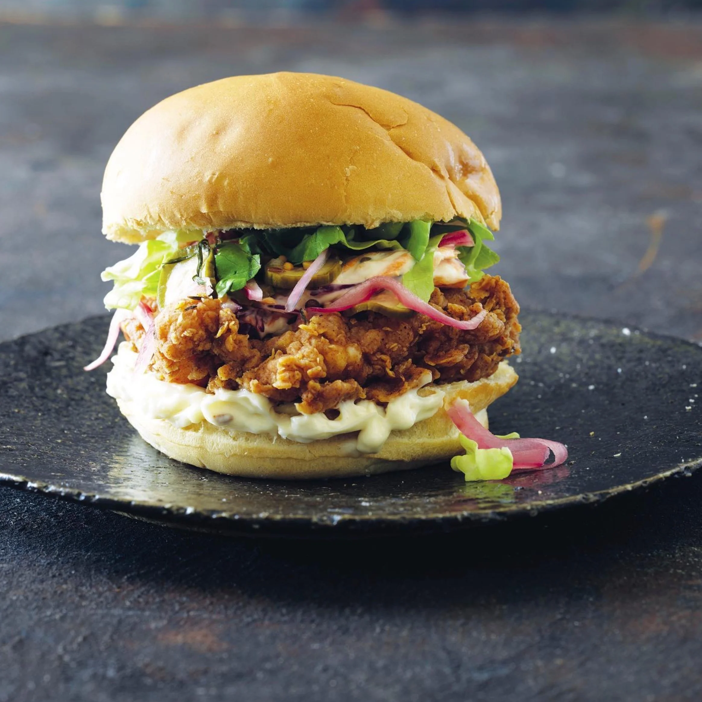 Close-up of a fried chicken sandwich with lettuce, tomato, and mayonnaise on a bun, placed on a dark surface.