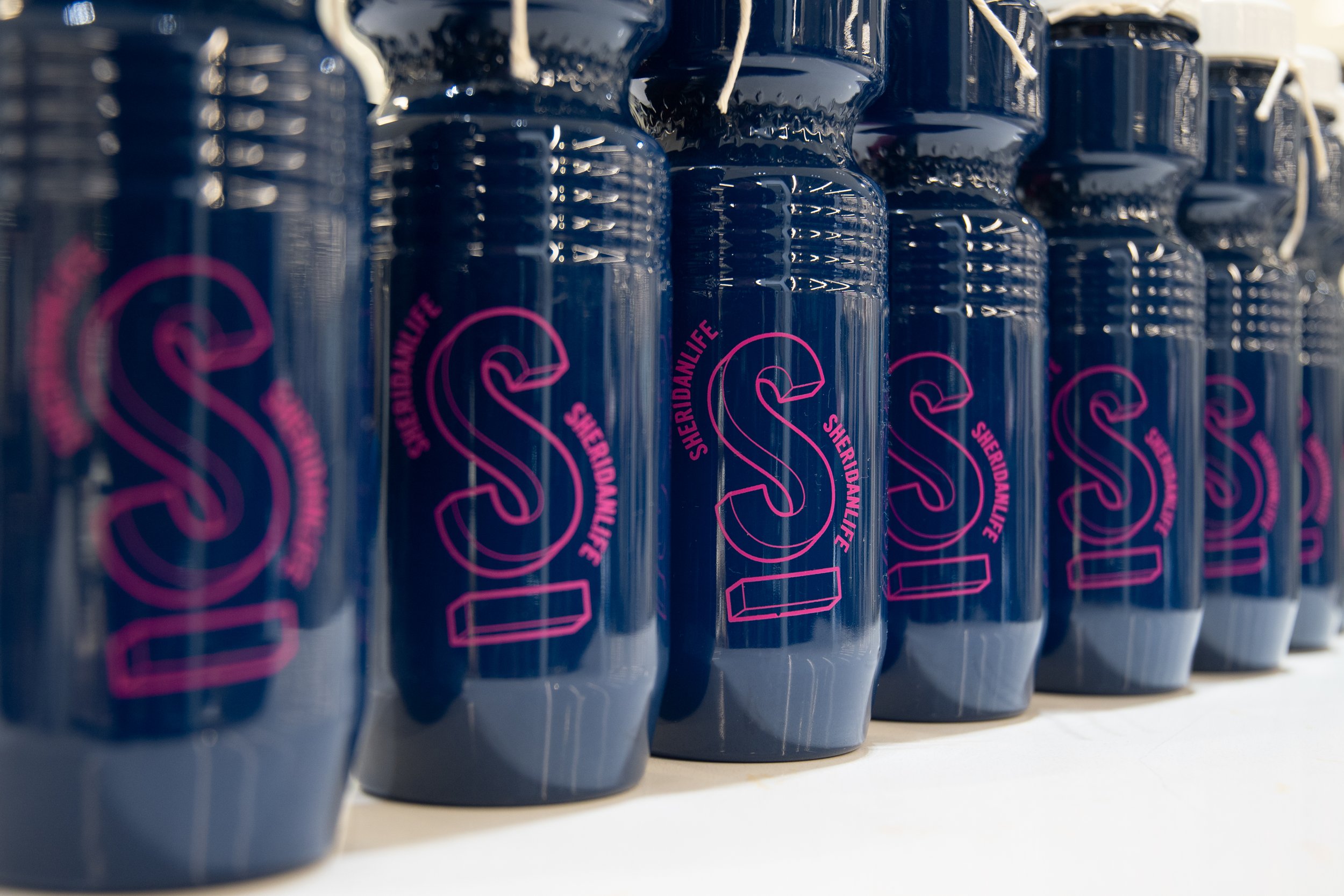  A row of navy water bottles in a diagonal line with giant magenta S’s on the side surrounded by Sheridan Life.  
