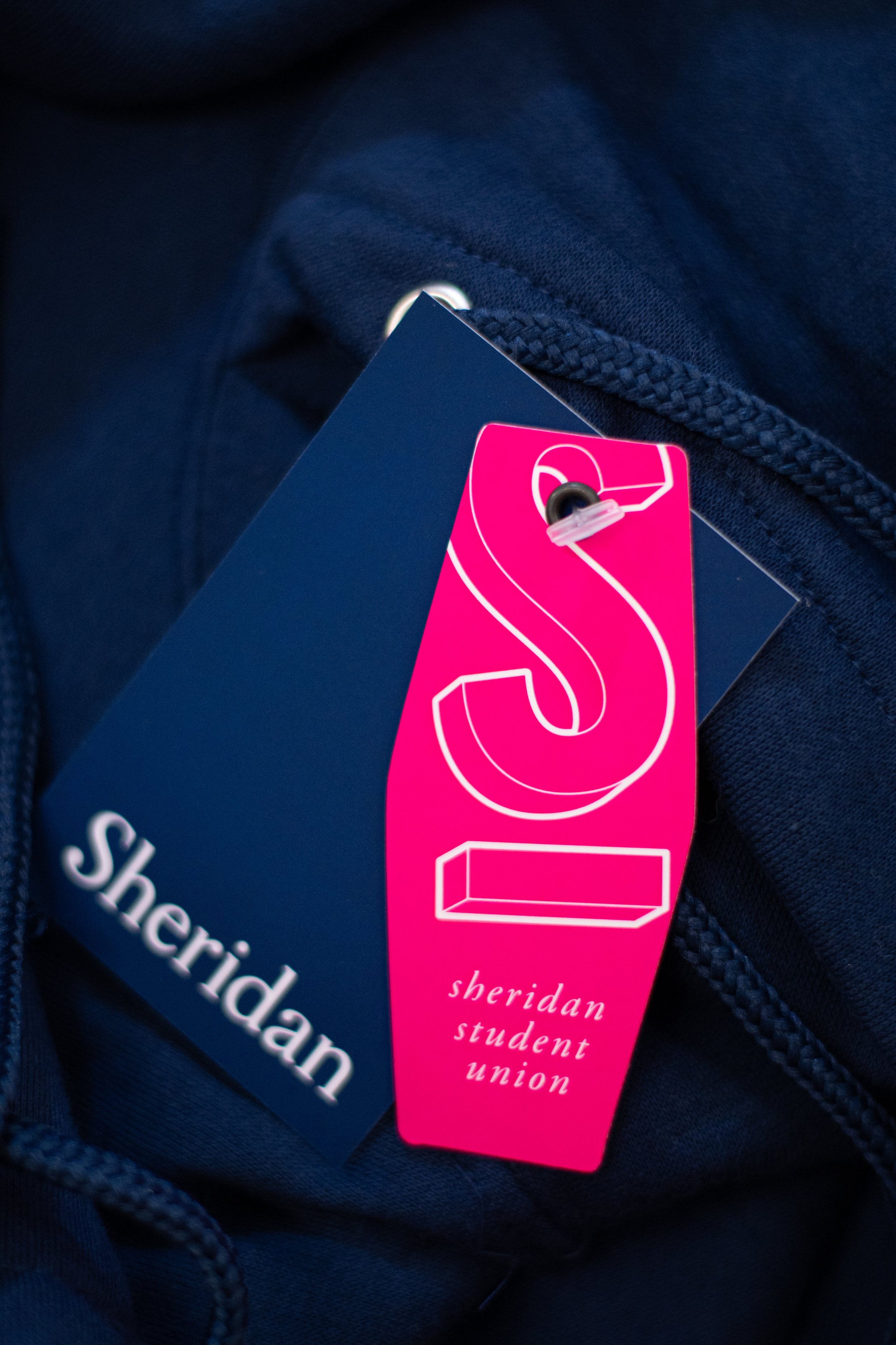  A magneta hang tag with an S at the top and a line underneath, Sheridan Student Union, attached to a blue Sheridan College tag, white Sheridan.  