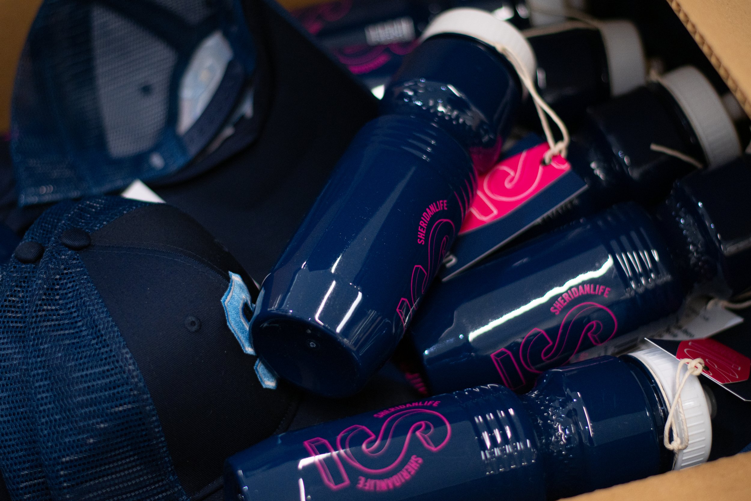  A cardboard box open with a collection of navy SSU water bottles  with white caps. On the bottles a “S” in magenta 