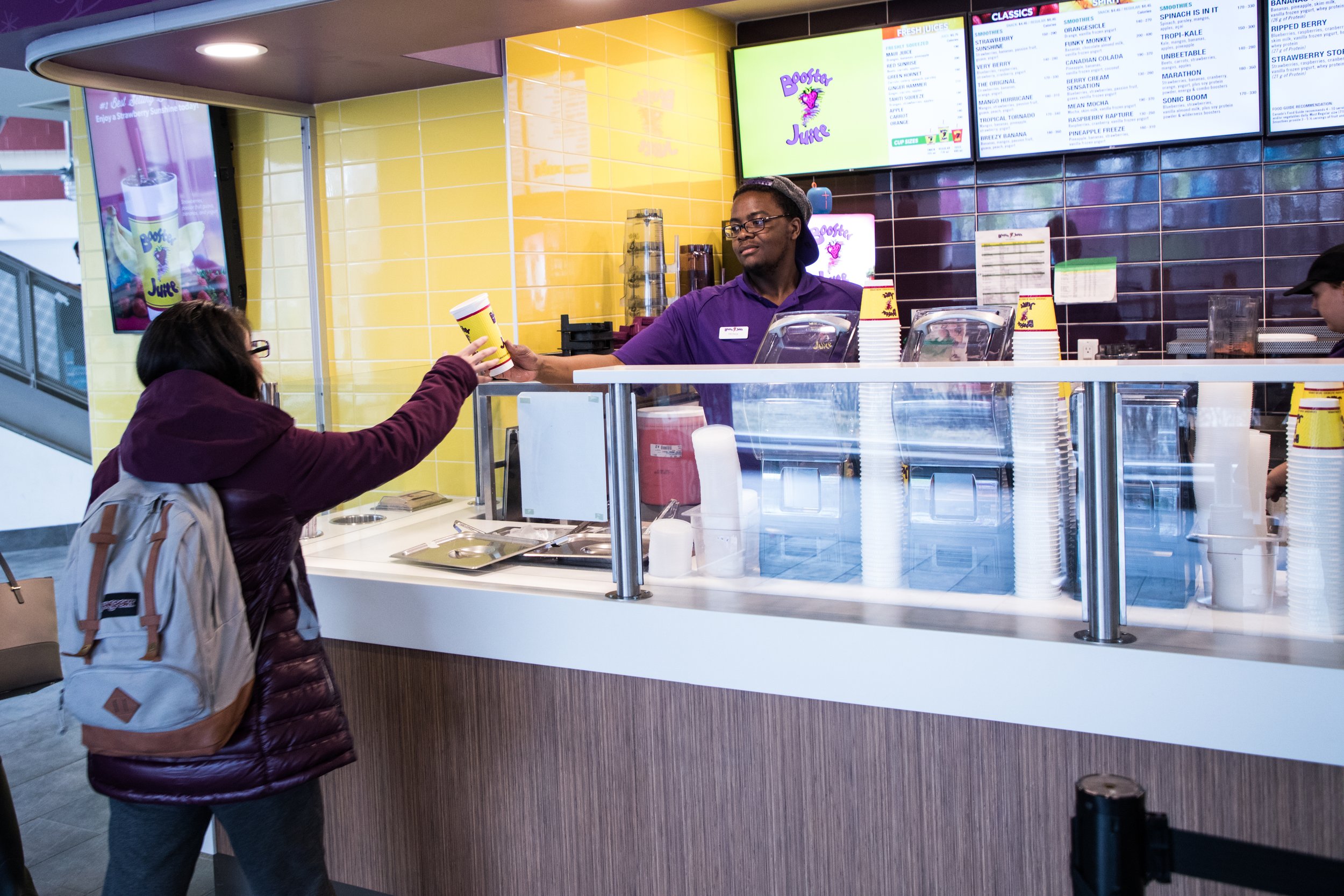  Booster Juice employee in a purple Booster Juice outfit, standing behind the desk, hands a student a Booster Juice drink.  
