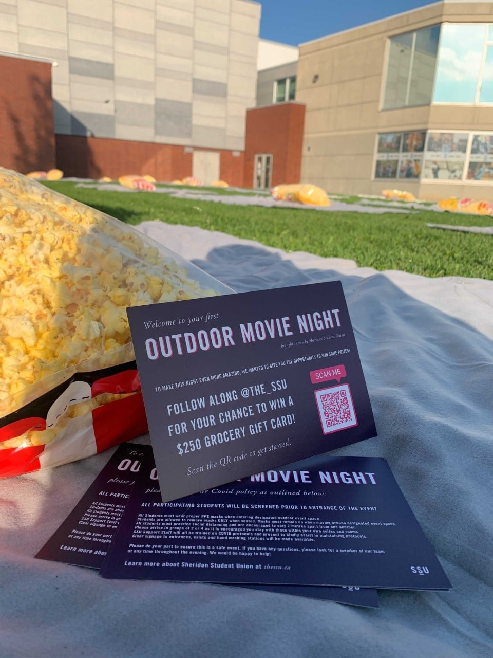  A postcard on a blank outside reads “Outdoor Movie Night” in front of a large bag of popcorn with the Sheridan College building in the background. 