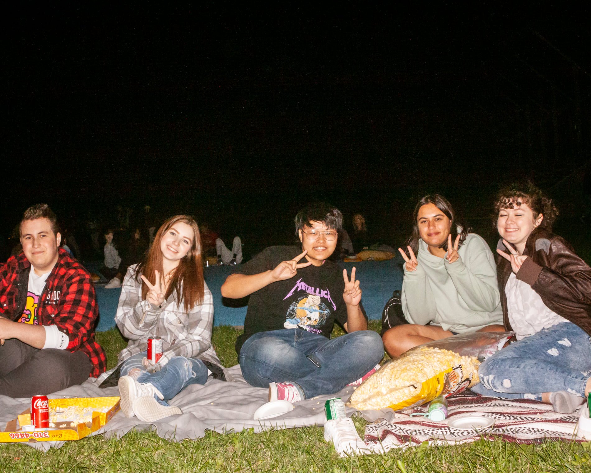 A group of 5 students sitting on the grass smiling surrounded by large popcorn bags at night.  