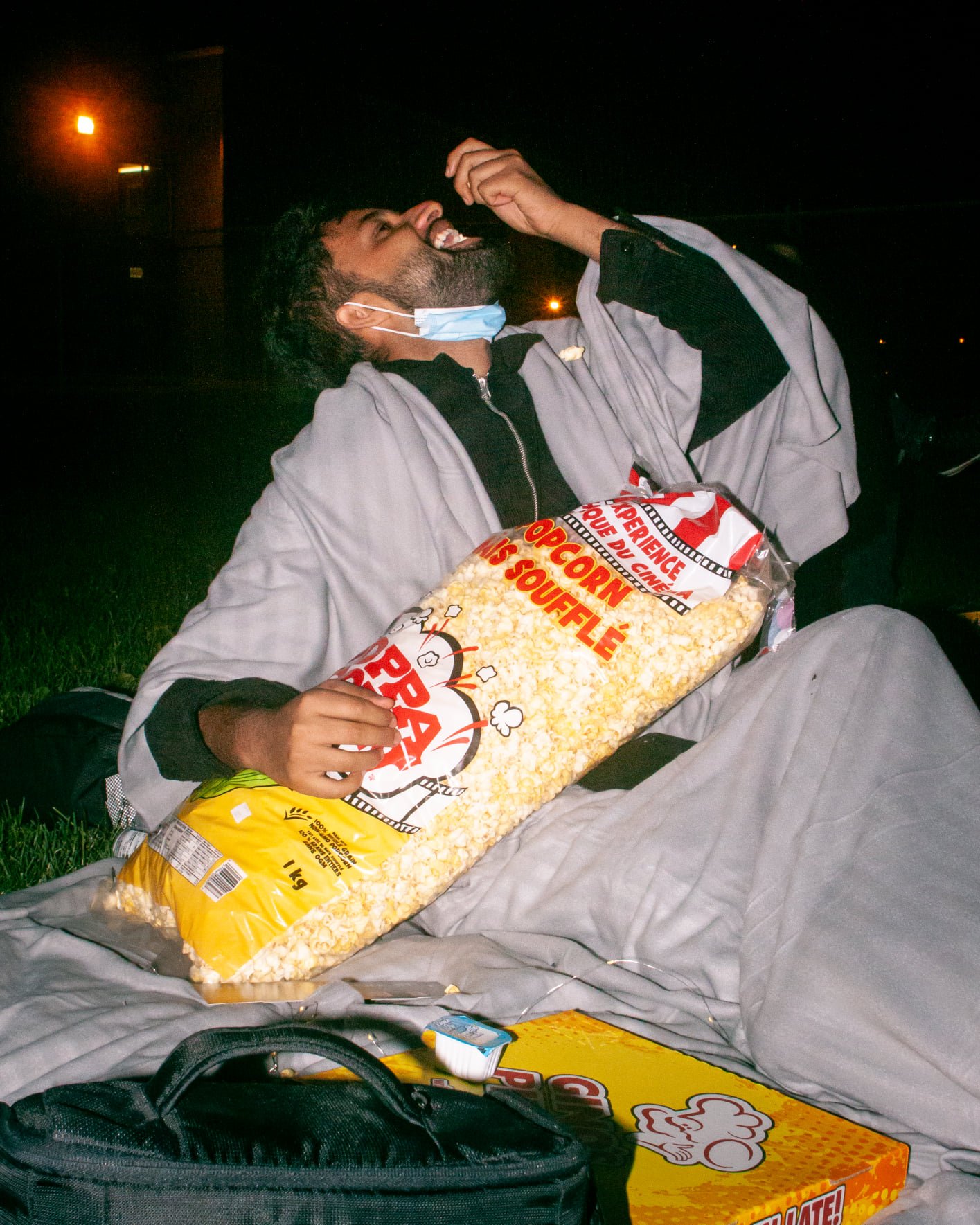  A side-profile view of a student holding a large popcorn bag, with a hand full of popcorn in the alternate hand with their mouth slightly open and head tilted back. 