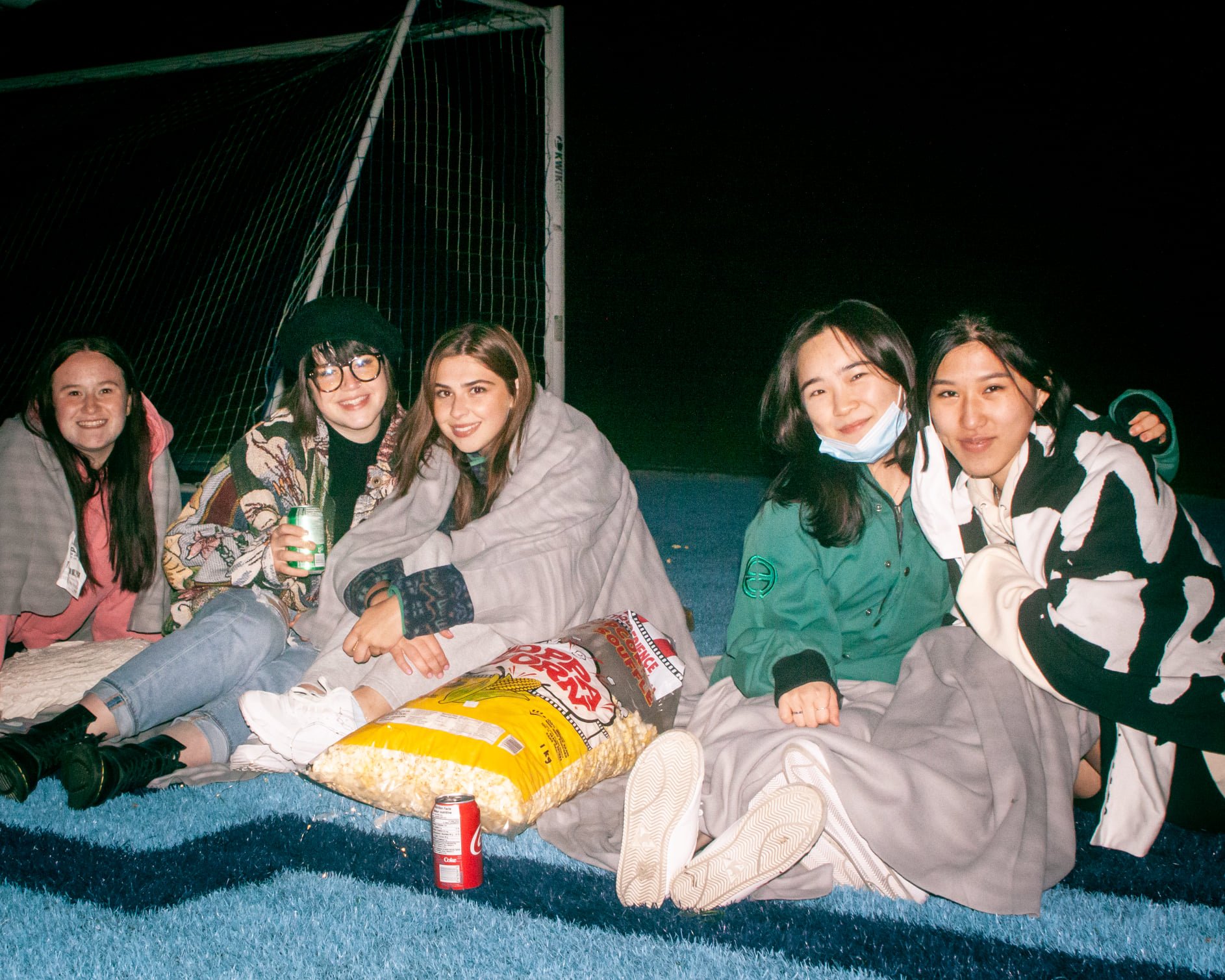  A  group of five students sitting on the grass smiling at night. A large popcorn bag in the middle.  