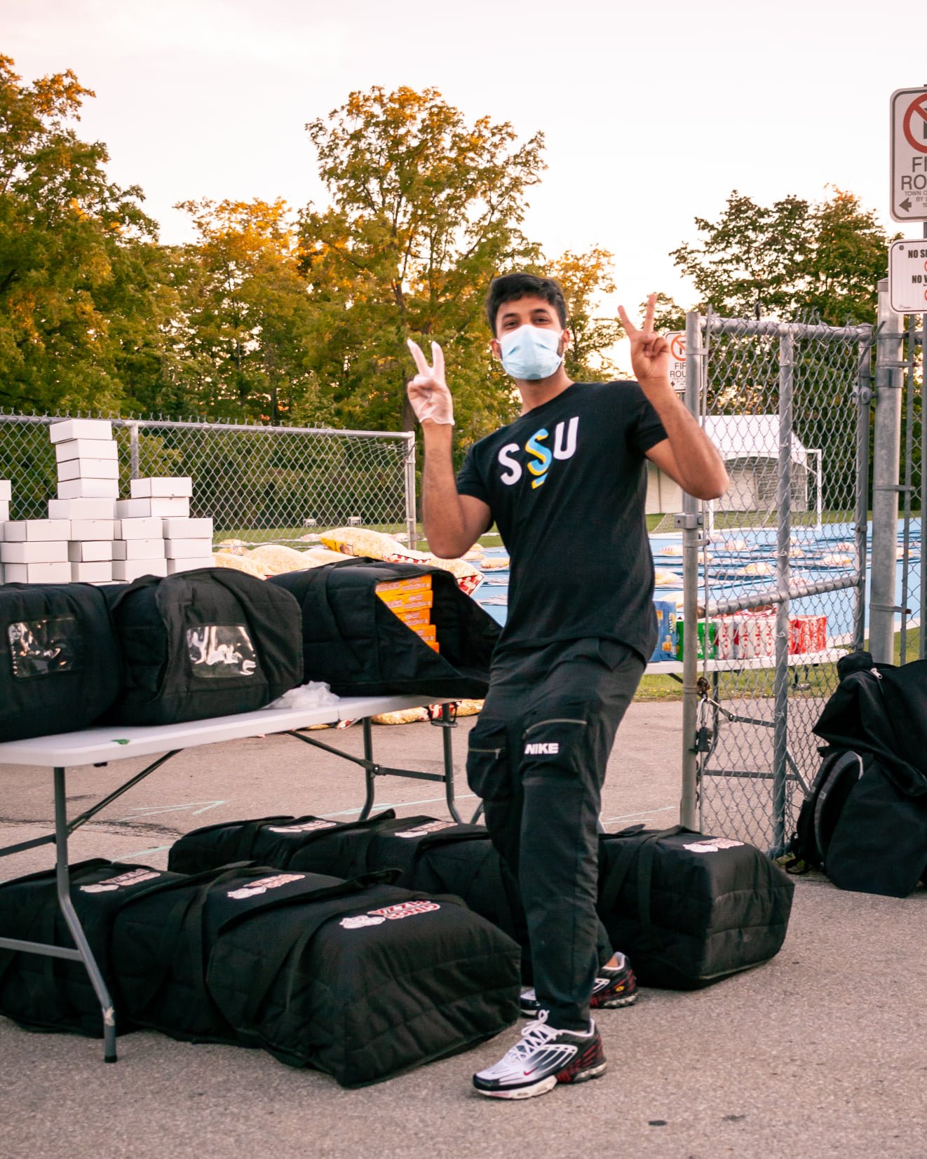  Student Life Coordinator standing at a table with food and pop cans in the distance, wearing a black SSU t-shirt, black pants and mask, giving a peace sign. 