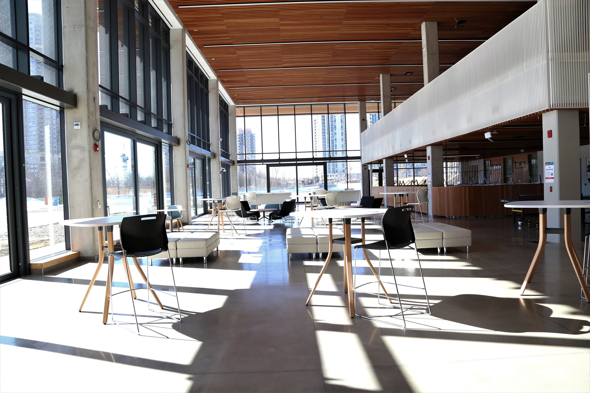  Inside the HMC Student Centre Atrium. Wooden panel ceiling, with glass windows all around, modern individual chairs and round tables in the centre of the floor.  