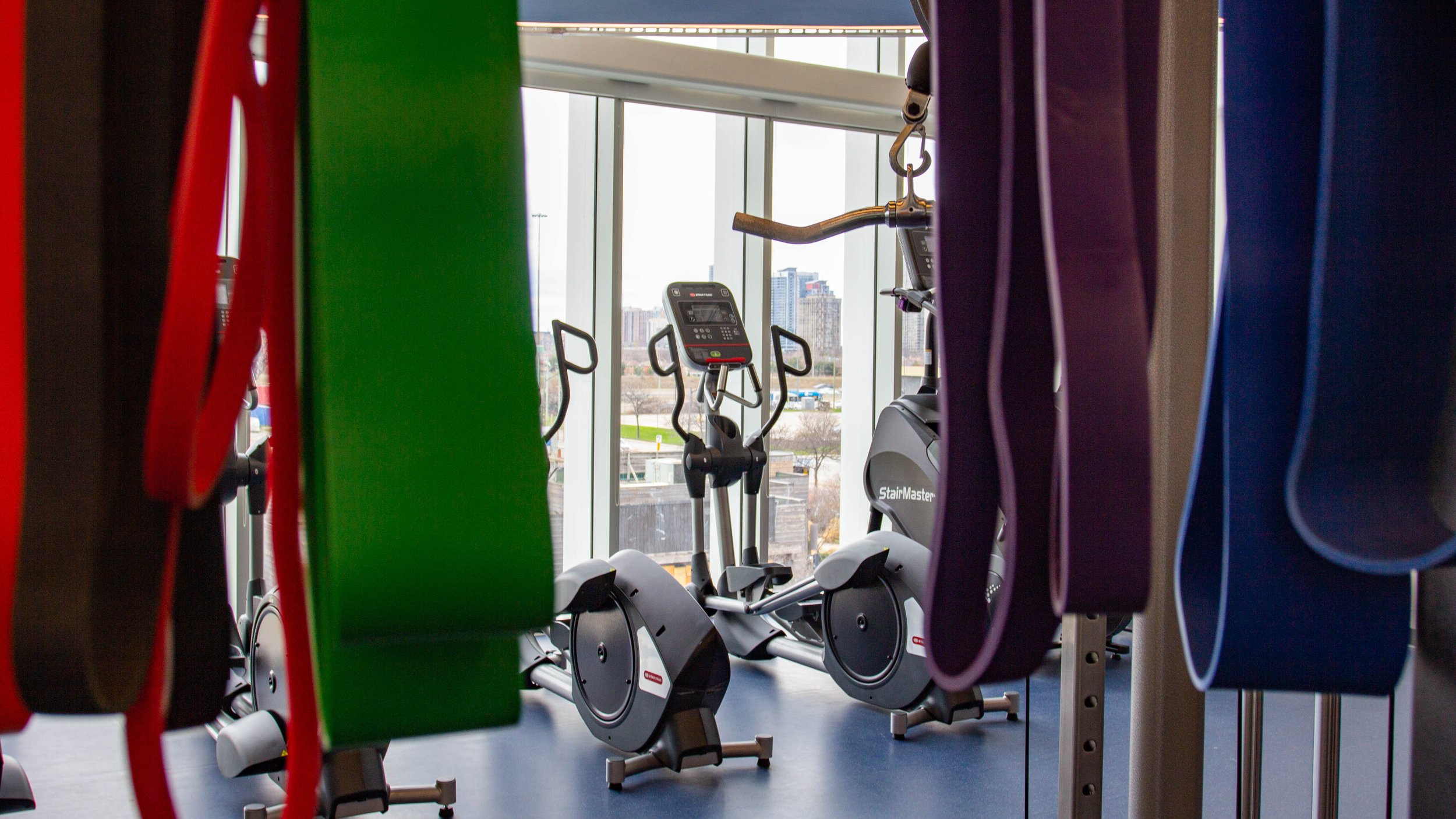  Close up of 4 large resistance bands in red, green, purple and blue hanging with a gap in the middle. In the distance, through the gap, you can see the elliptical machine. 