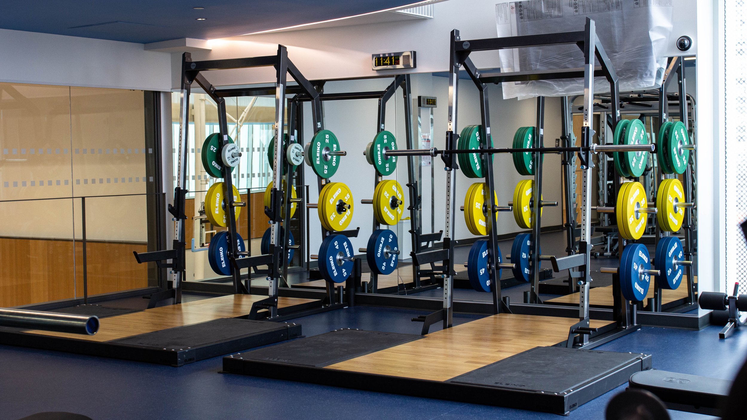  A bar of three sets of weights: on the bottom blue weights, then yellow and then green. Underneath a navy mat with mirrors behind the weights.  