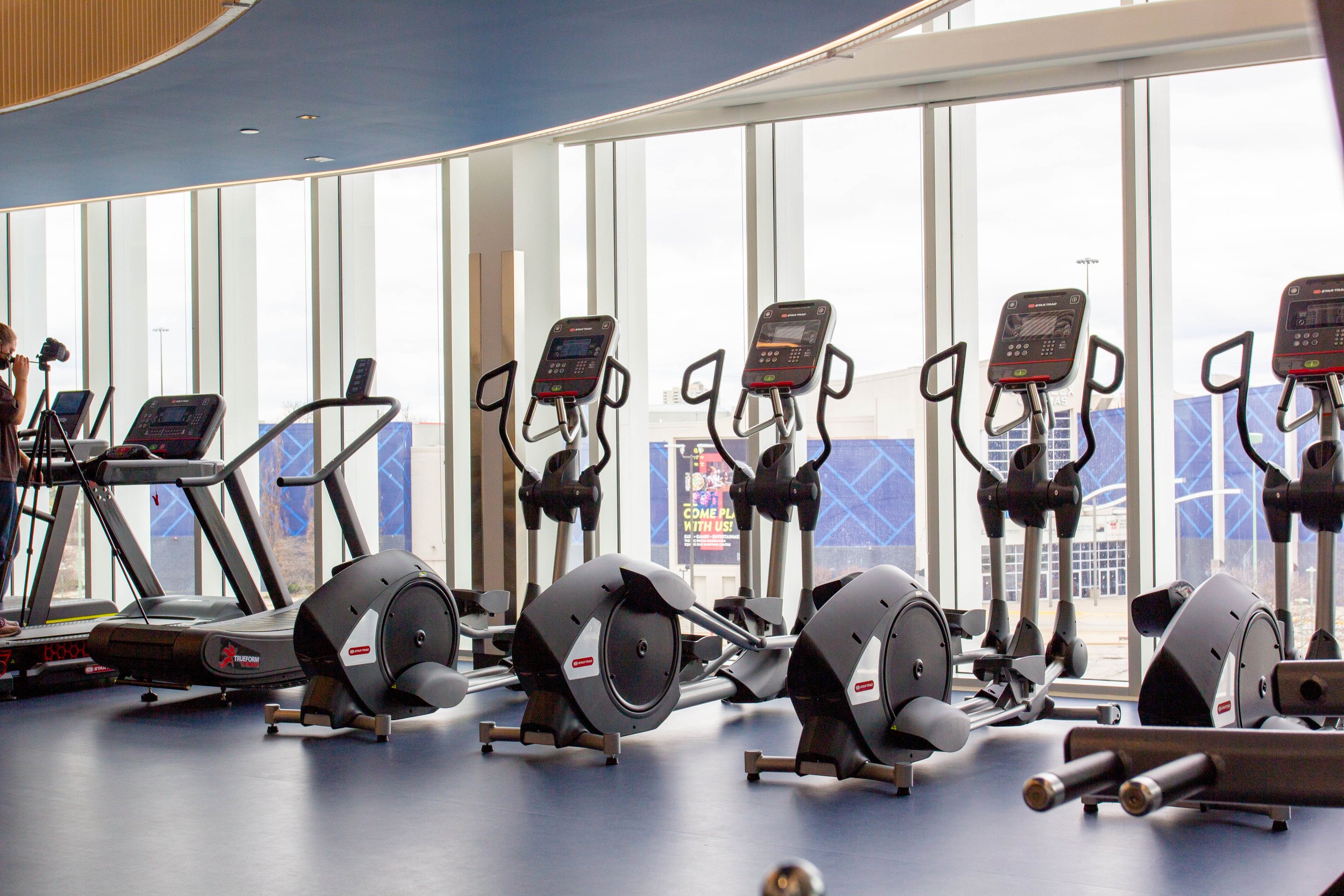  A row of elliptical machines and treadmills facing a glass window on a navy blue floor.  