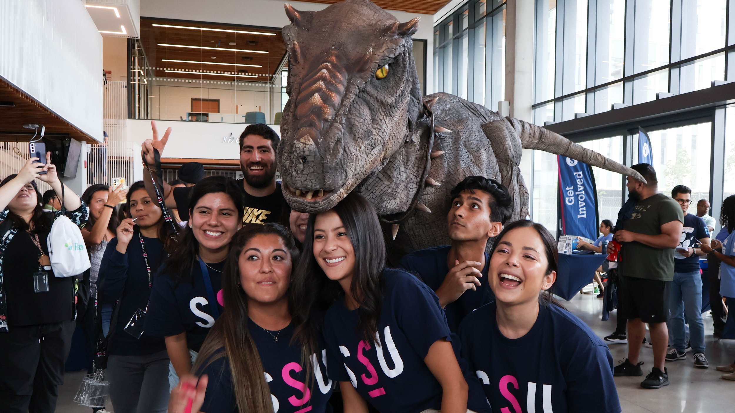  A group of students in navy SSU t-shirts smiling with a T-Rex above their heads at the HMC Student Centre.  