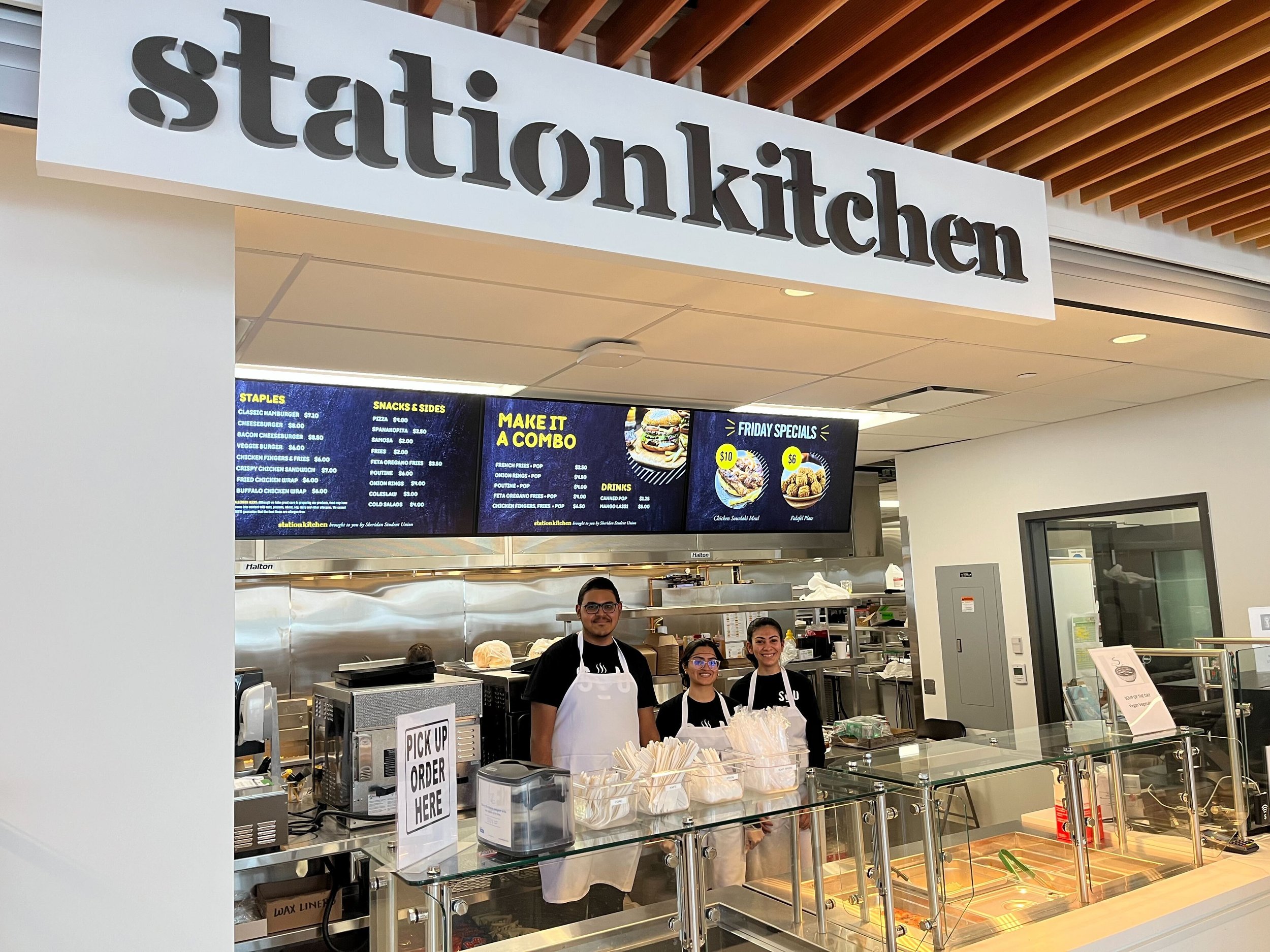  Station Kitchen sign underneath wooden panels. A digital menu above three students in black SSU shirts and a white apron standing inside the kitchen. 