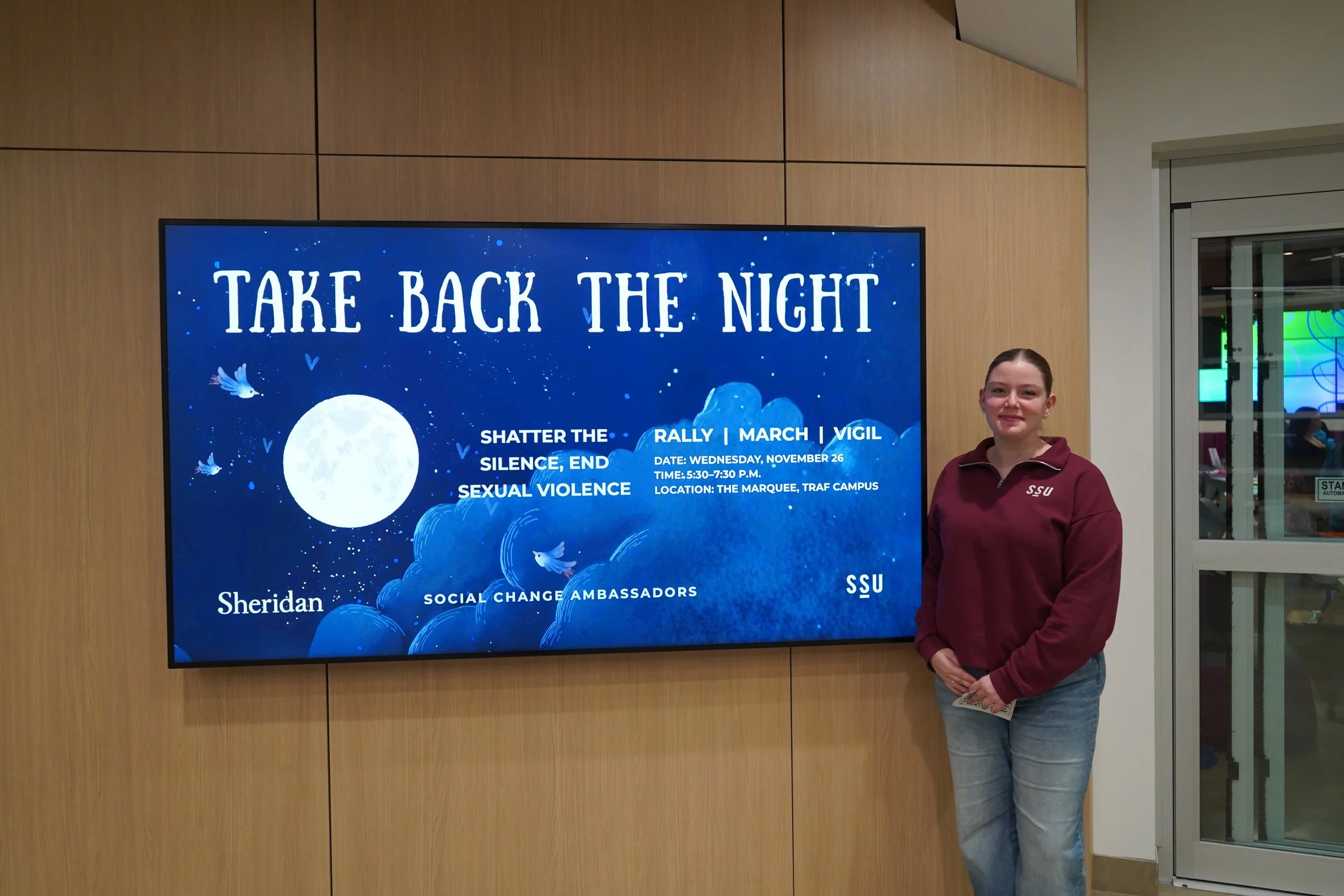 A girl standing next to the TV with the Take Back The Night visuals on it,